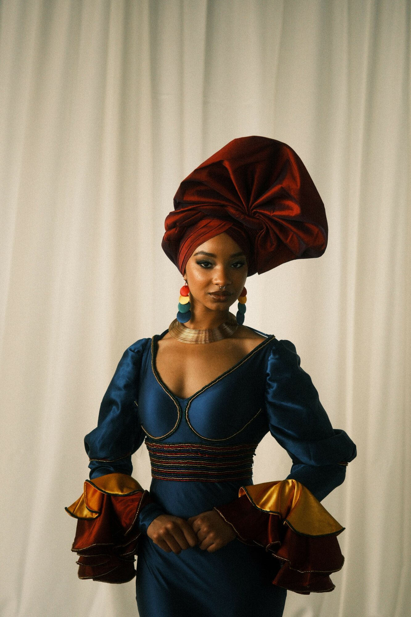 Woman in colorful traditional attire with headwrap, standing against a plain background.