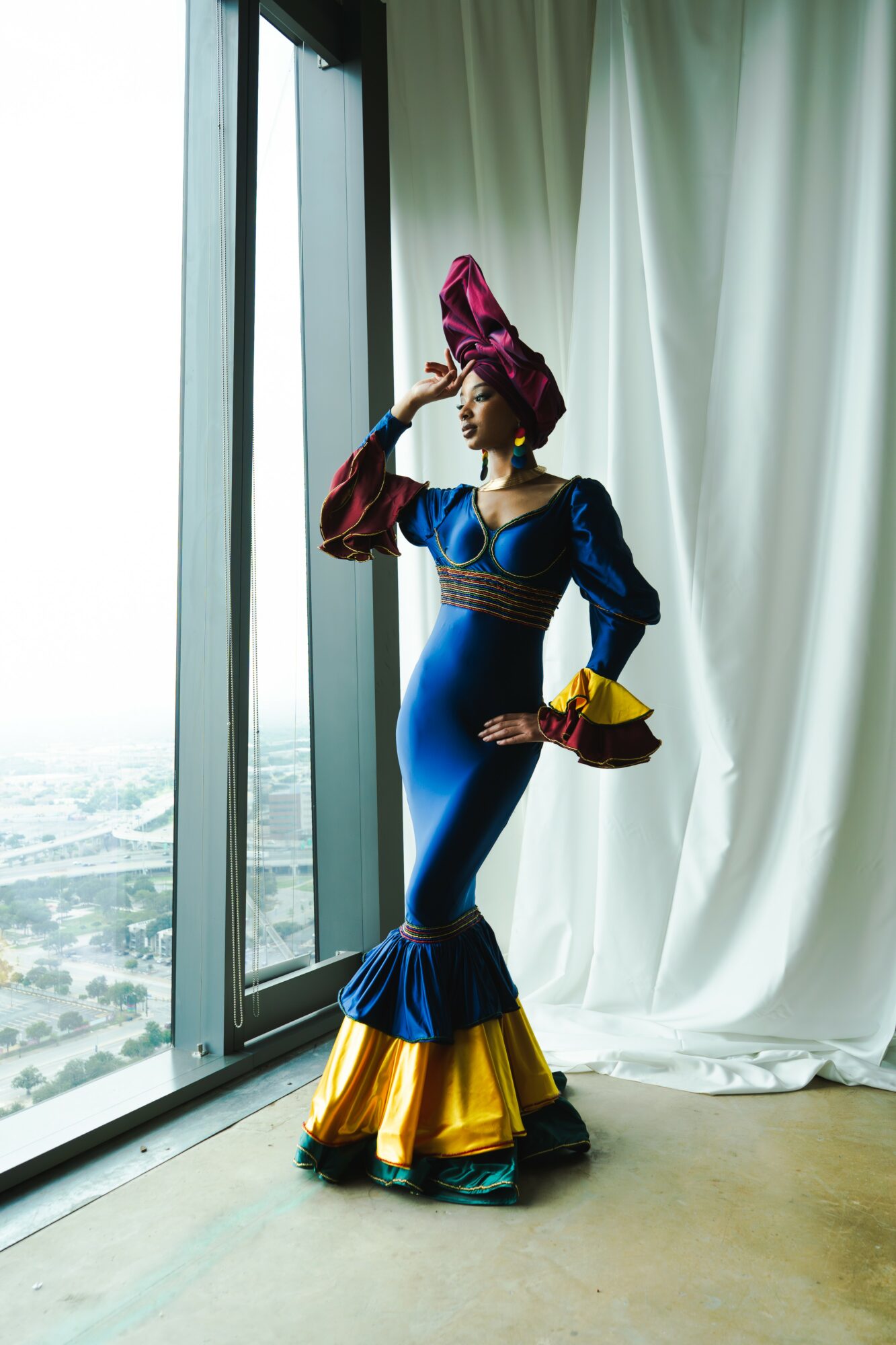 Person in colorful costume standing by window with curtains, looking outside, holding a hat, with city view outside.