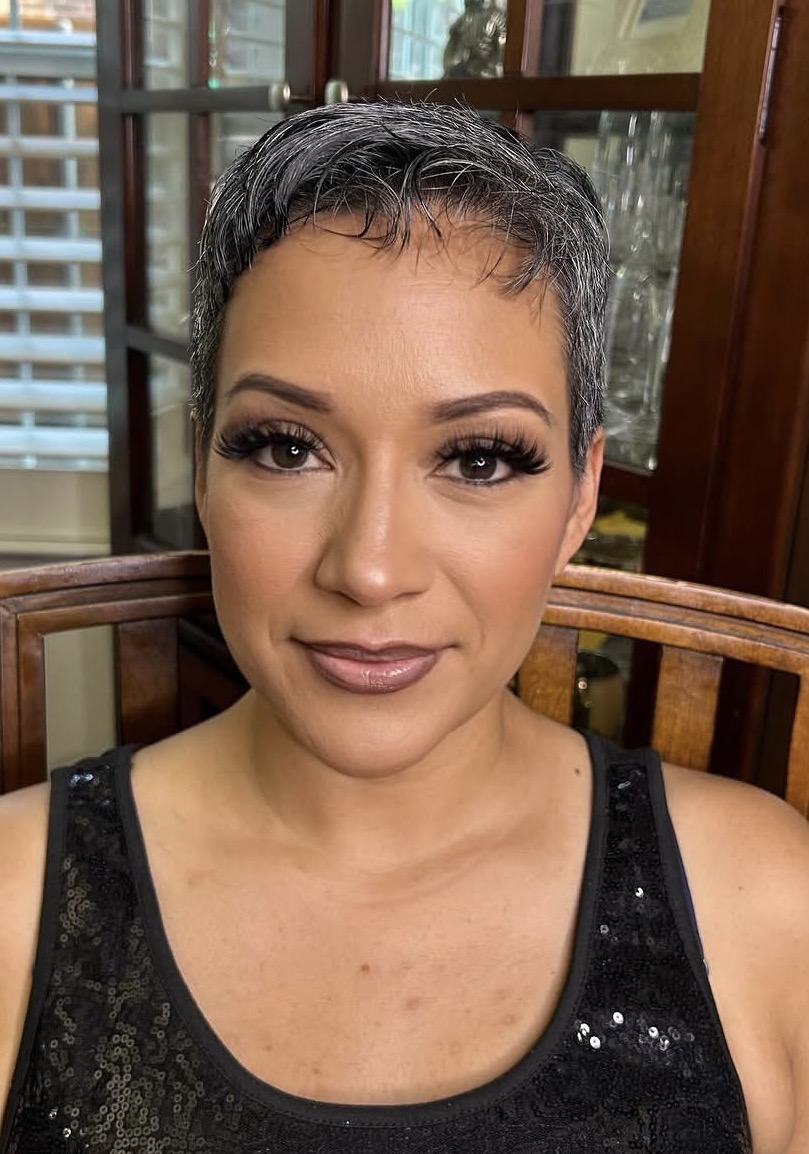 Woman with short gray hair and makeup, wearing a black sequined top, sitting indoors near a window.