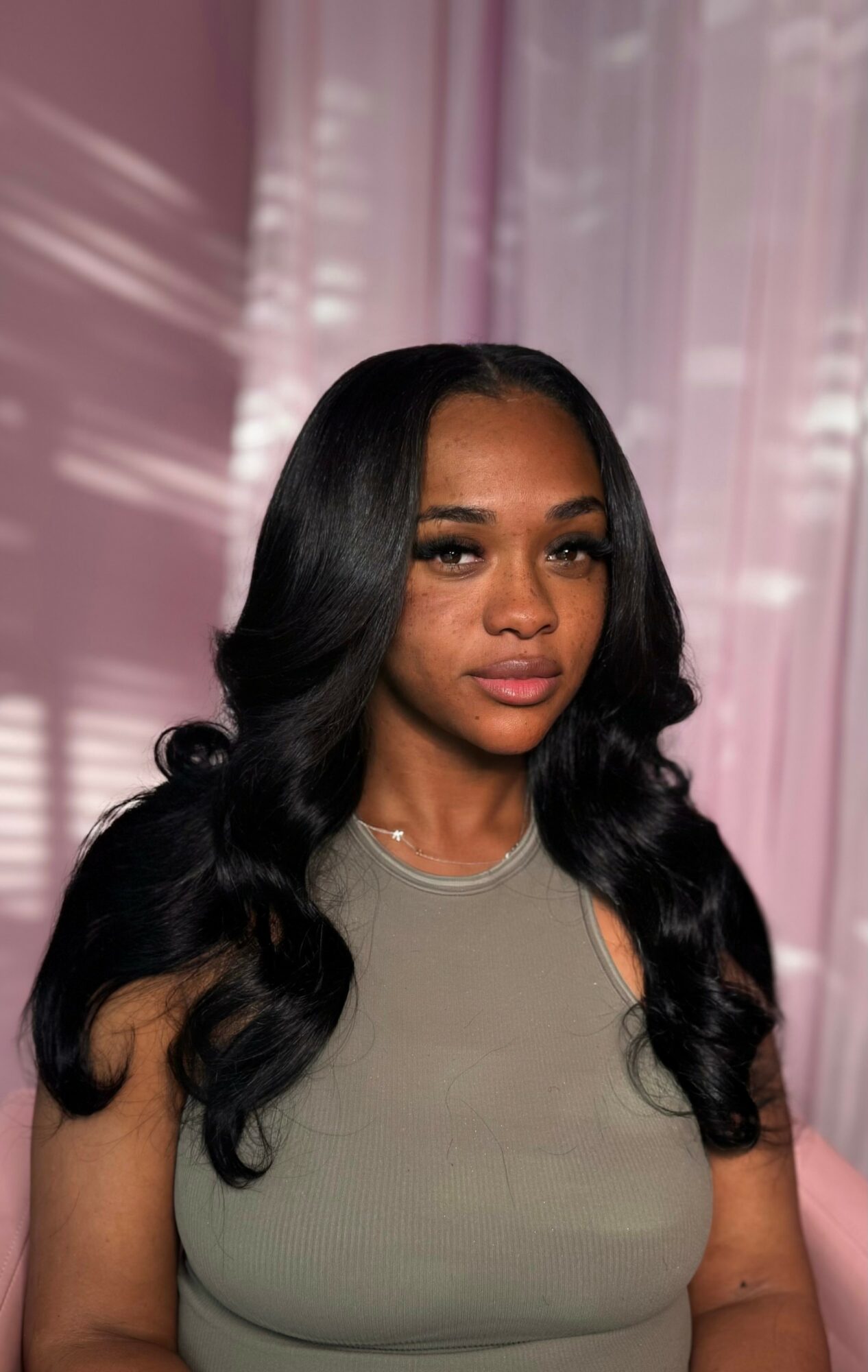 A woman with long, wavy black hair and a neutral expression stands in front of pink curtains with light patterns.