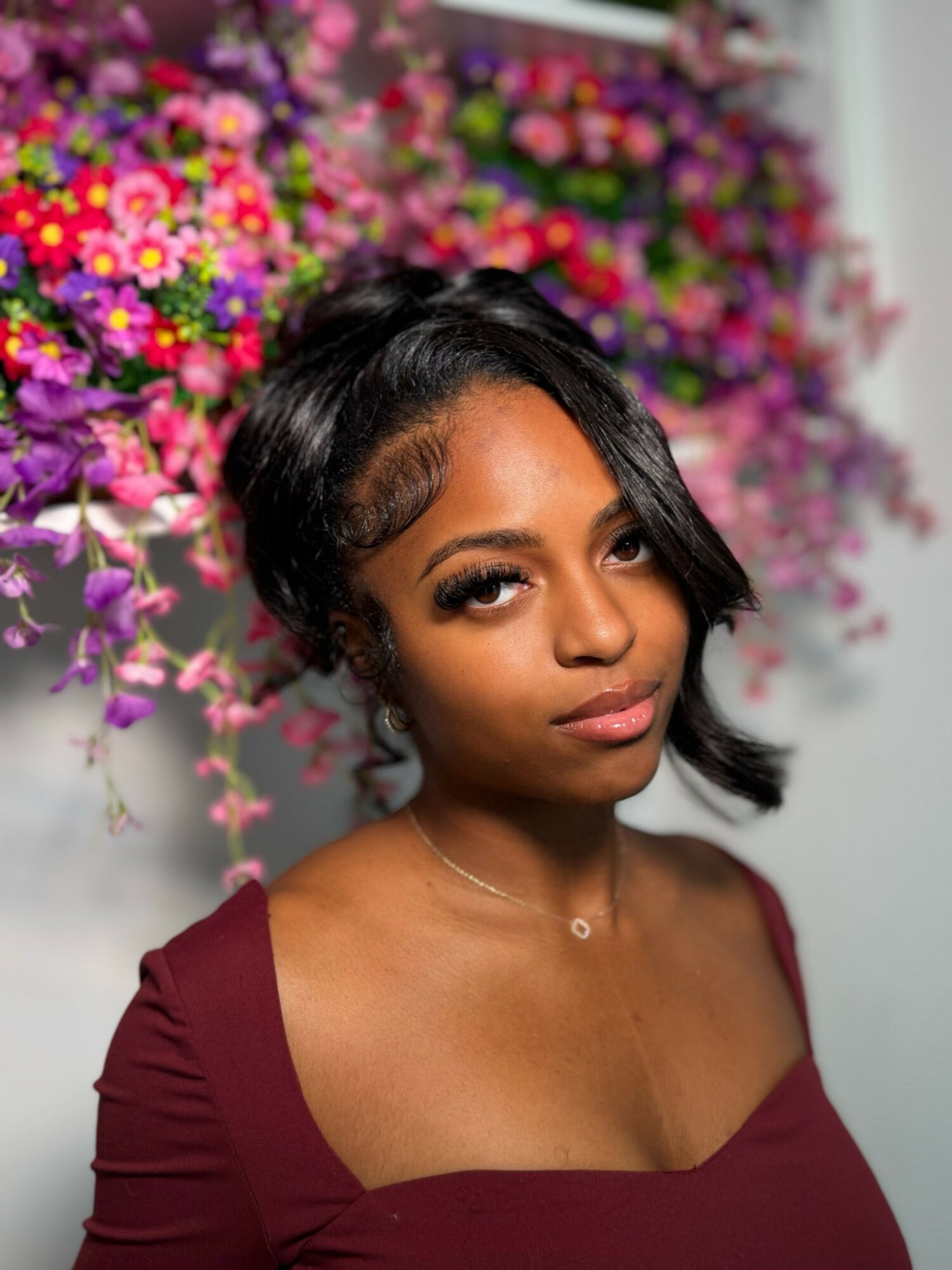 A woman with short black hair and makeup, wearing a burgundy top, stands in front of a colorful flower arrangement.