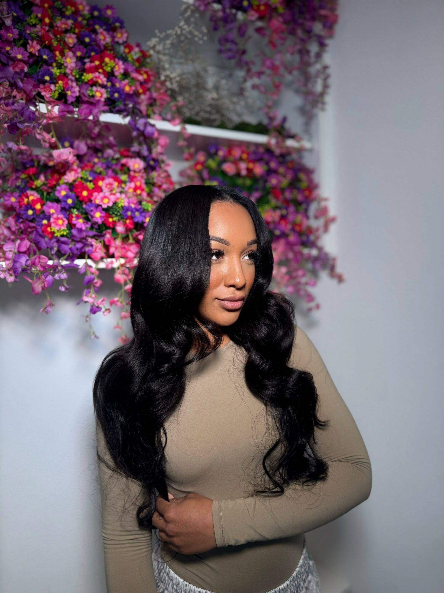 Woman with long black wavy hair standing in front of colorful flower arrangements, wearing a beige top, looking to the side.