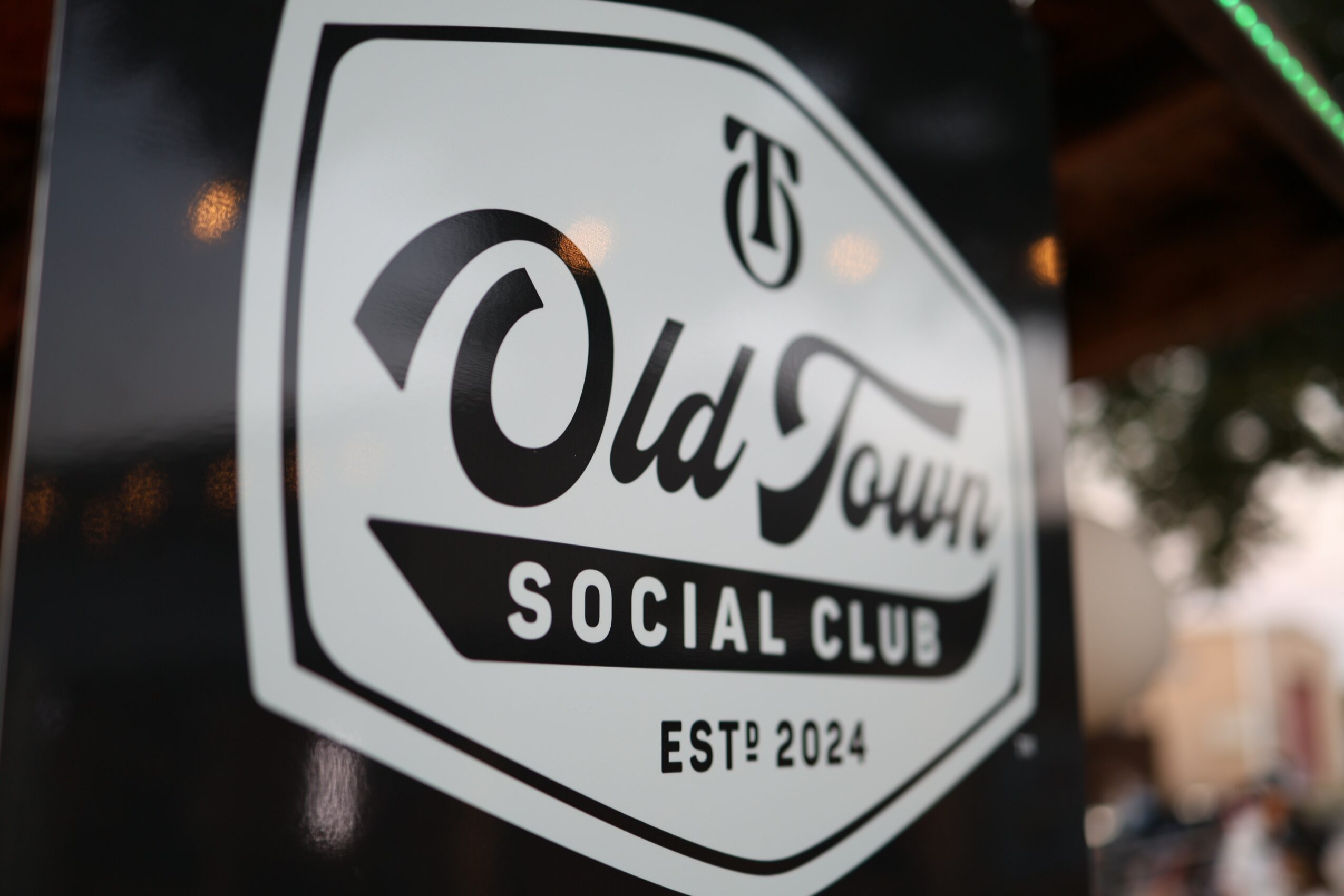 Sign for Old Town Social Club with logo and establishment year 2024, outdoors setting, blurred background.