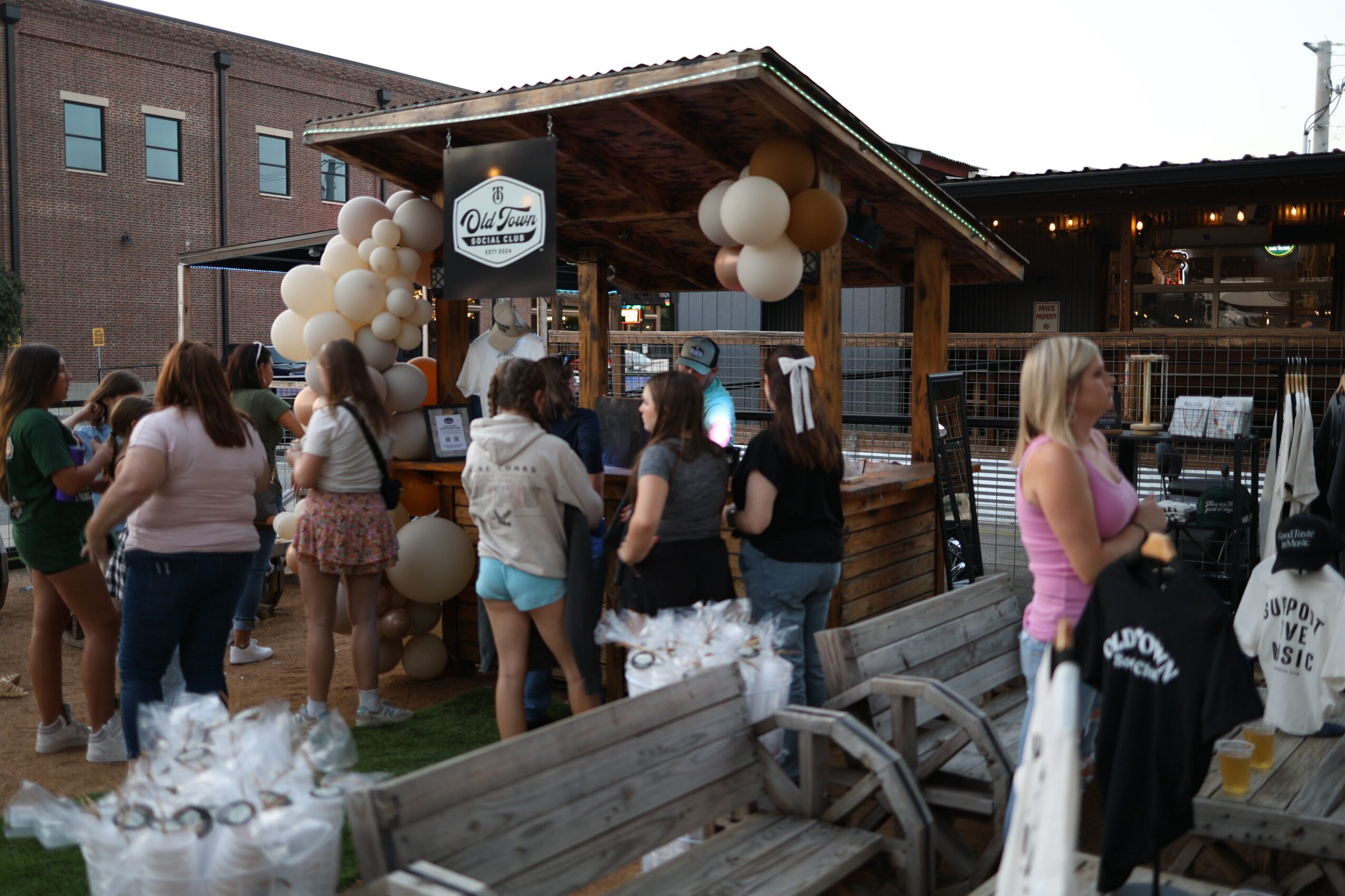 Group of people gathered outdoors near a wooden bar decorated with balloons, with benches and tables nearby.