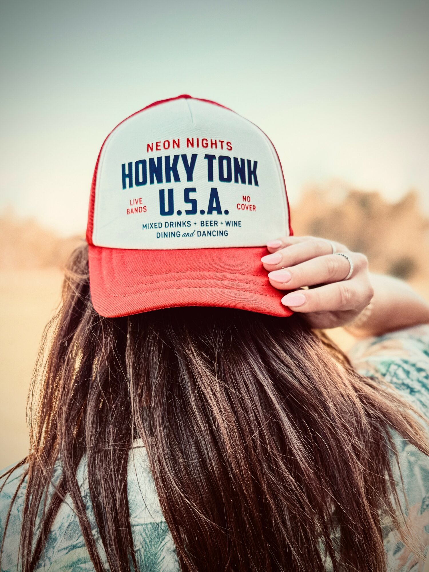 Person with long hair wearing a red and white trucker hat, holding it with one hand, outdoors with a blurred background.