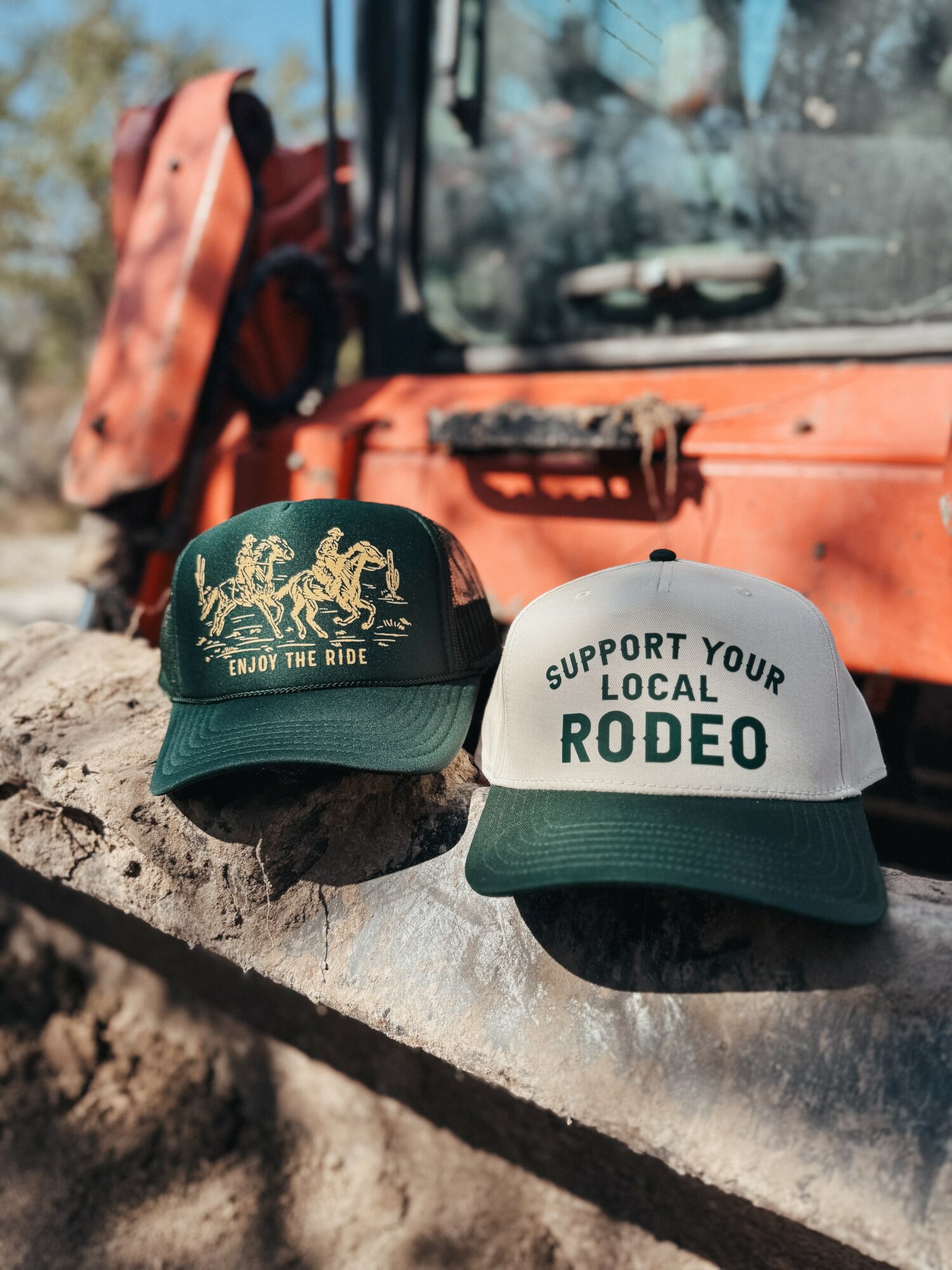 Two baseball caps, one green with a tiger graphic and the other white with text supporting local rodeo, placed on a concrete ledge.