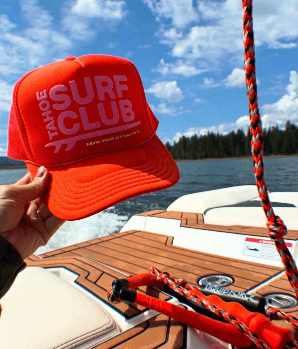 Hand holding a red cap with 'TAHOE SURF CLUB' text, boat deck, water, and sky in background.