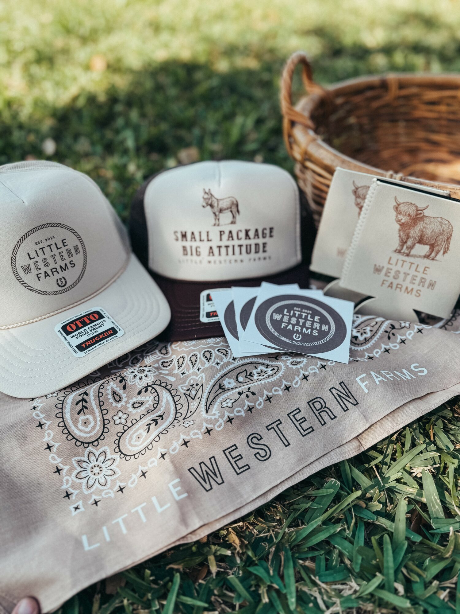 White cap, small package with text, stickers, and a basket on grass with a Little Western Farms cloth, outdoors.