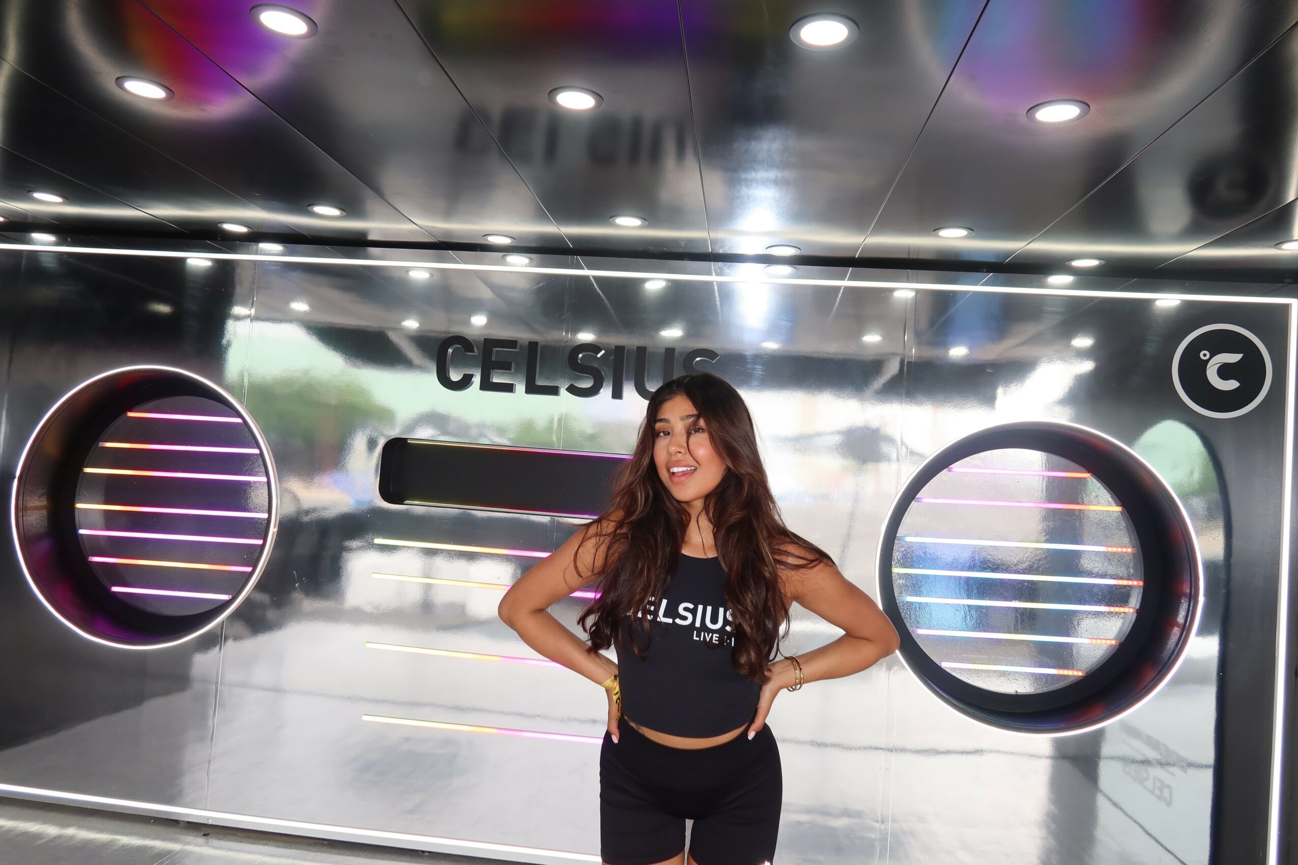 Young woman with long hair smiling, standing in front of a futuristic display with circular lights, hands on hips.
