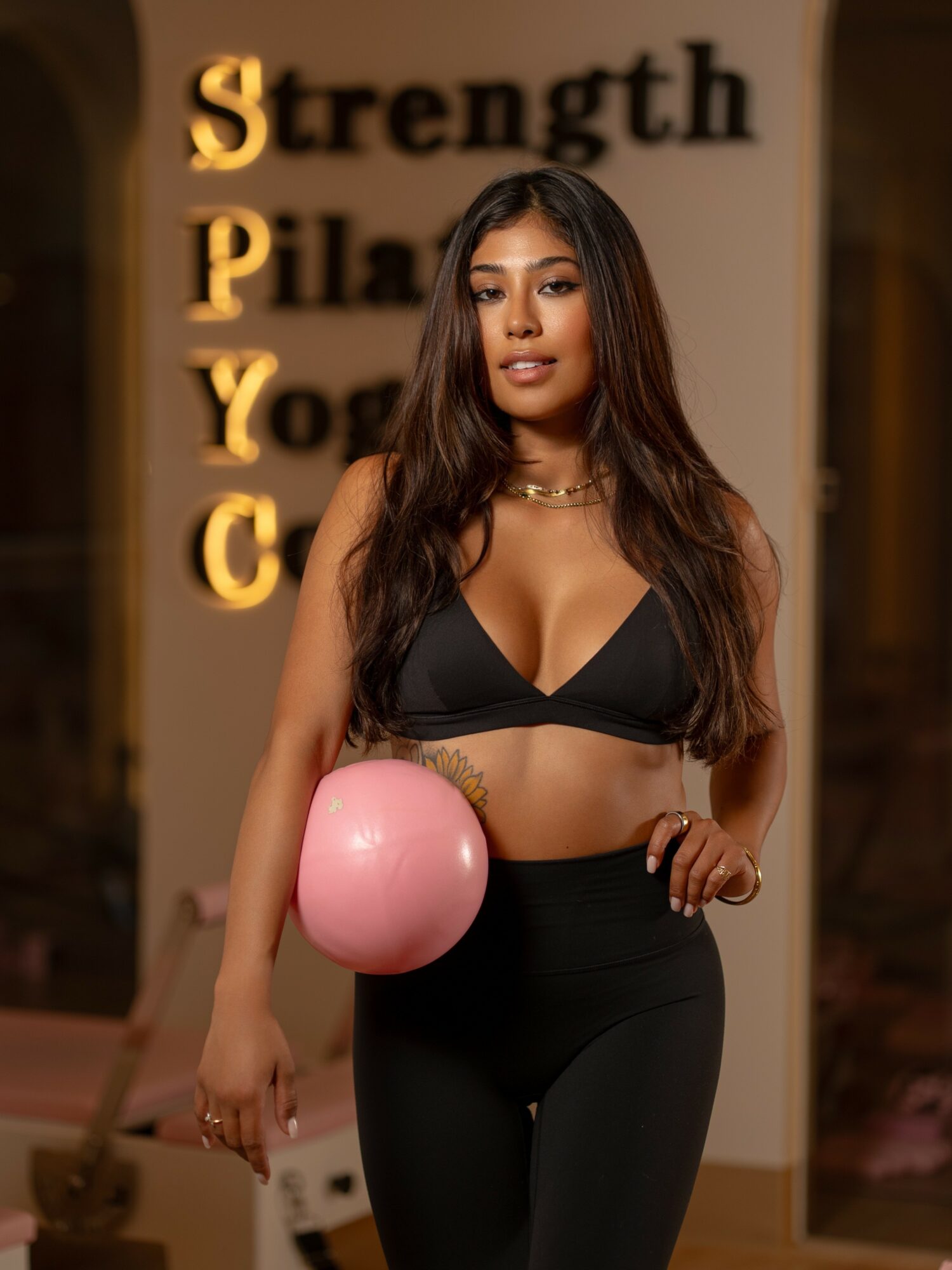 Young woman in black sports bra and leggings holding a pink balloon, standing indoors with a fitness-themed background.