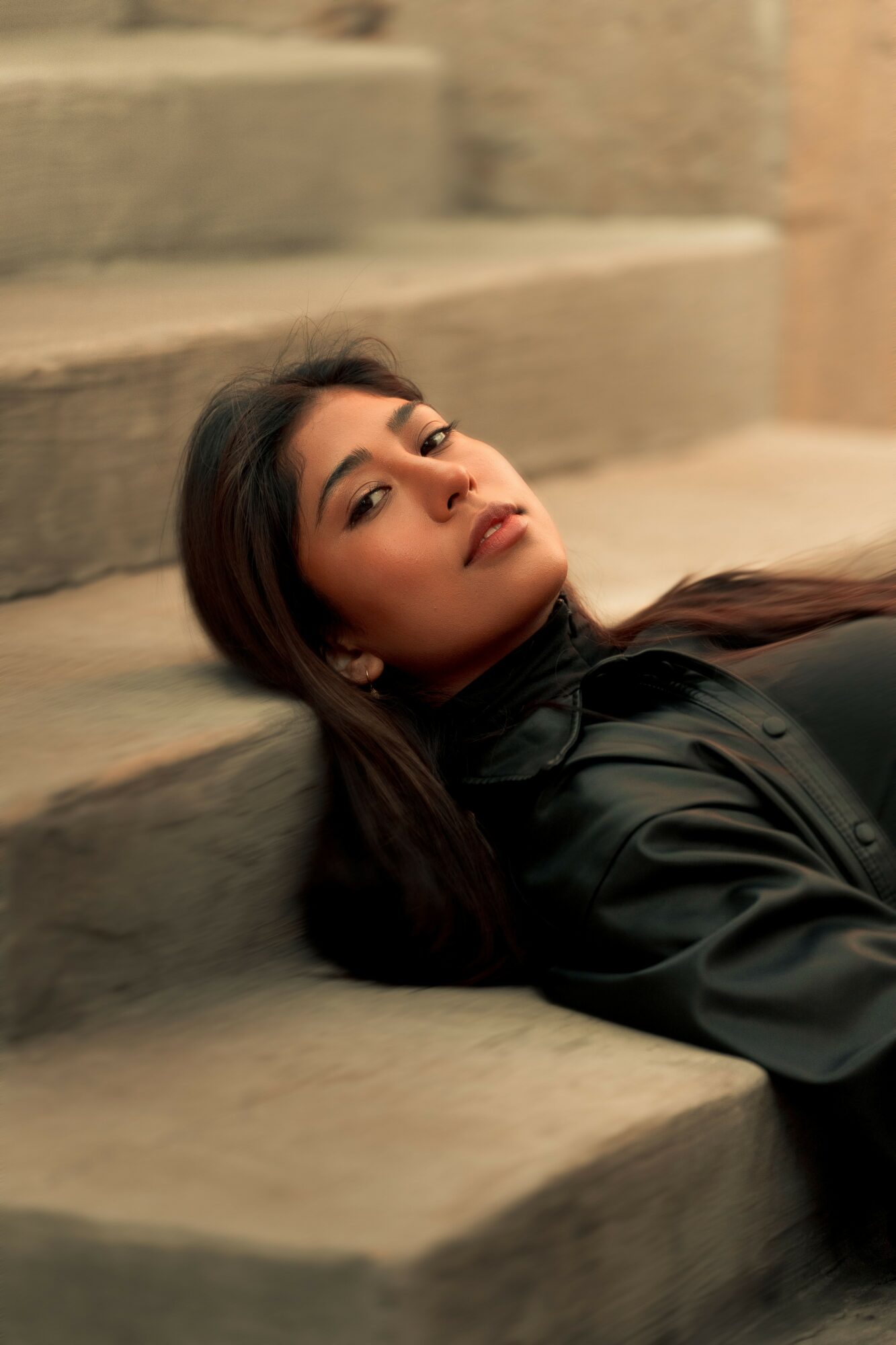 Woman with long dark hair lying on stairs, looking at camera, wearing a black jacket, neutral background.