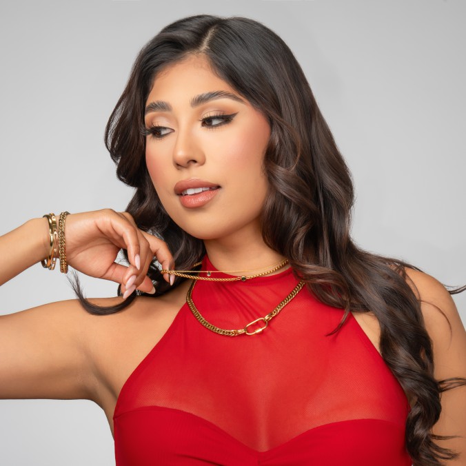 Woman with long dark hair wearing a red sleeveless top and gold jewelry, holding a necklace near her face.