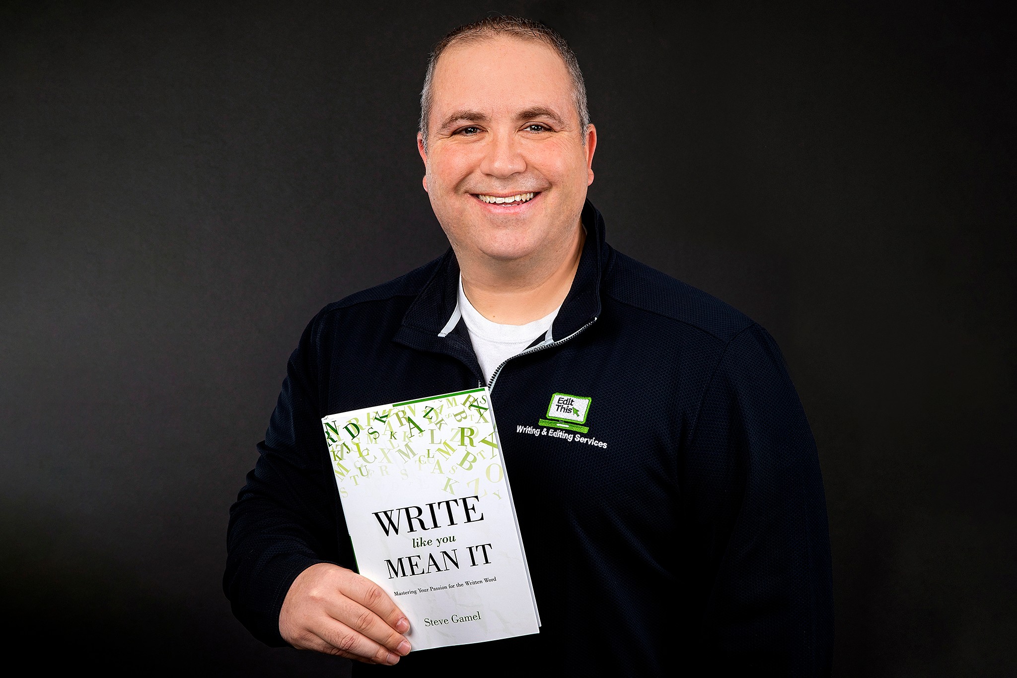 Man holding a book titled 'Write What You Mean It' smiling, wearing a dark jacket, against a dark background.