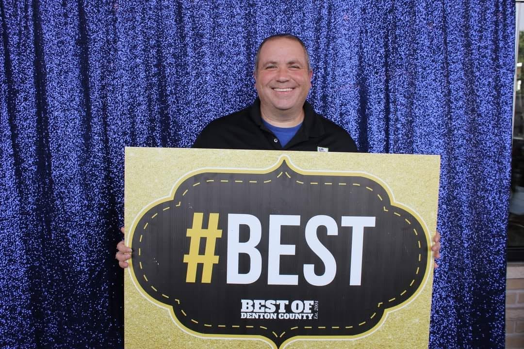 Man holding a large sign with '#BEST' in front of a blue curtain background.