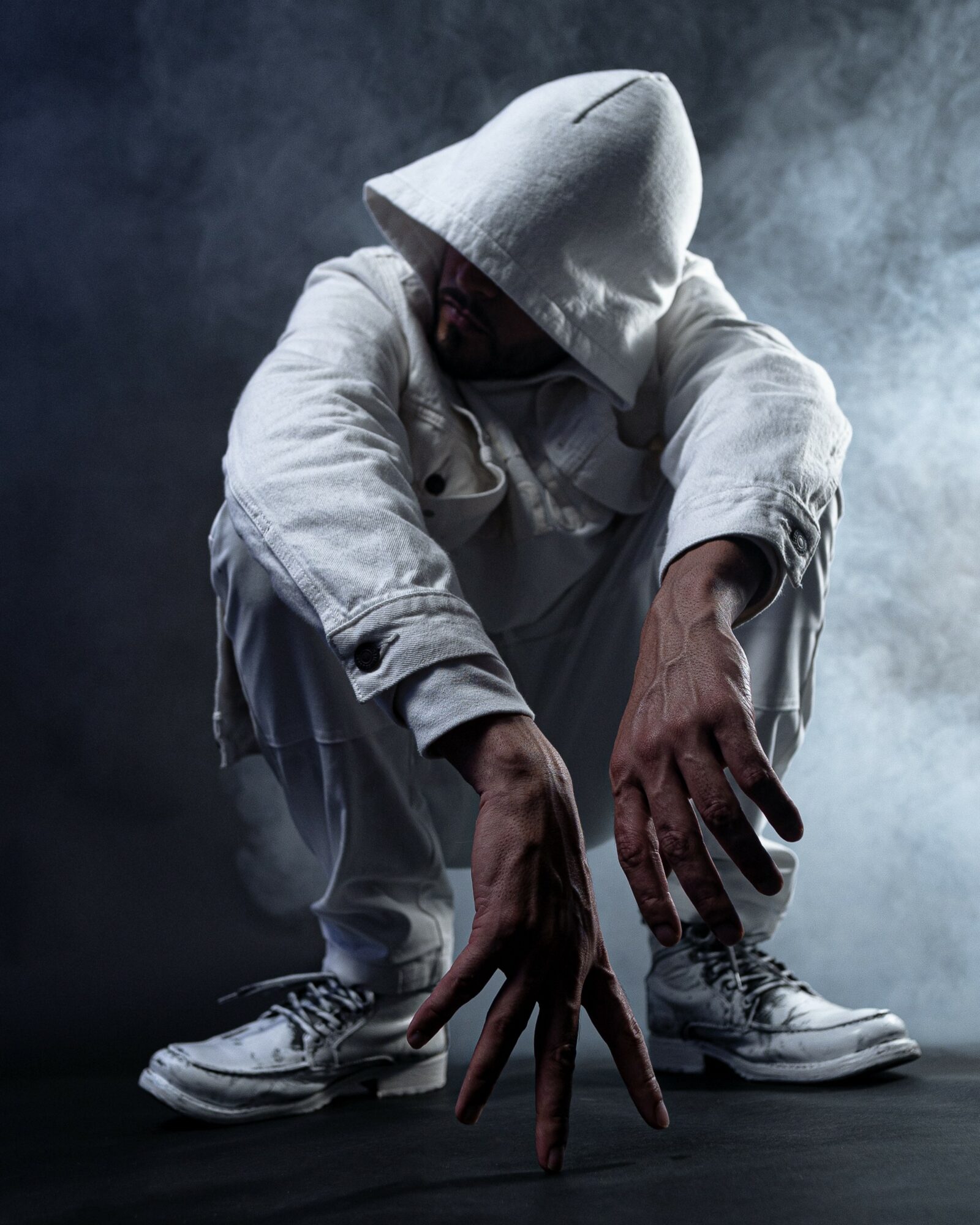 Person crouching with hood over face, hands extended forward, in a dark smoky environment.