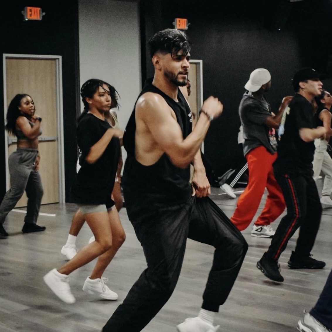 Group of people dancing in a studio, some with arms raised, wearing casual workout clothes, with a dark wall background.