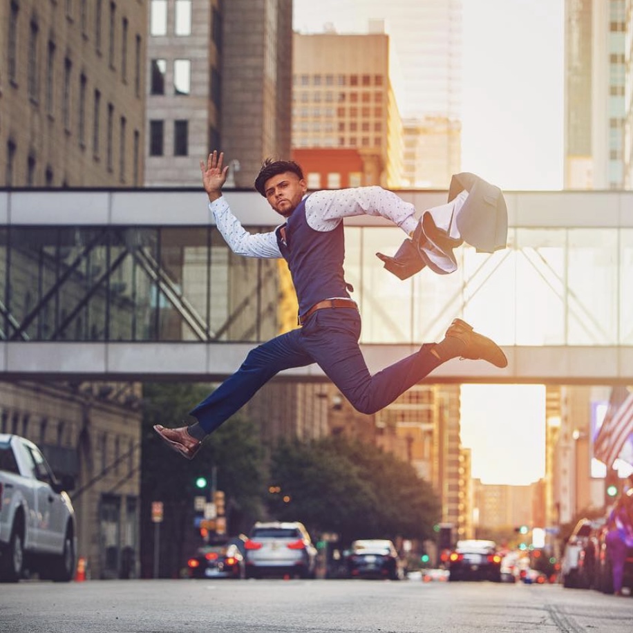 Man in business attire jumping in city street holding a briefcase, tall buildings in background, sunset lighting.