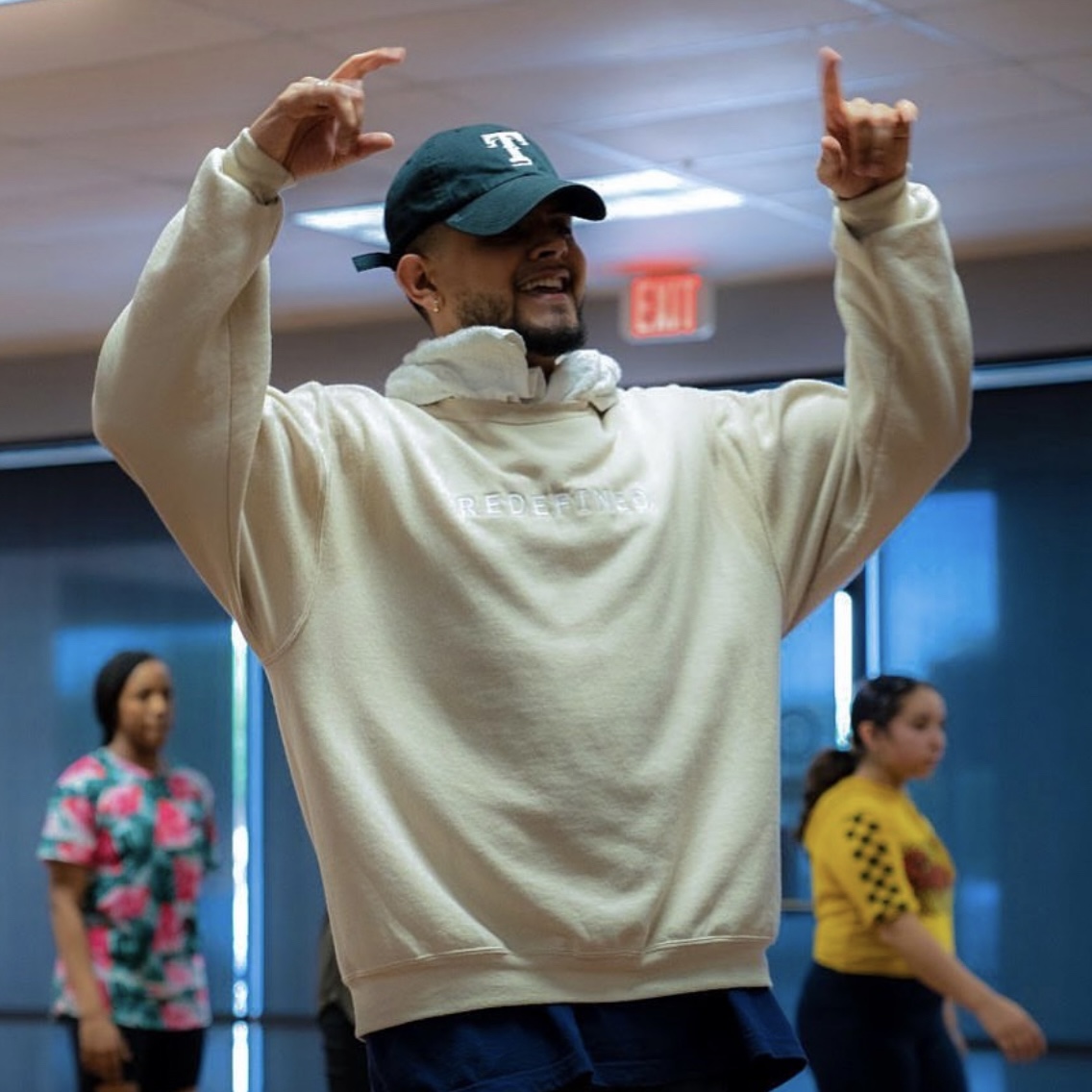Smiling man in a baseball cap and cream hoodie raising both hands indoors, with two women in colorful clothing in background.