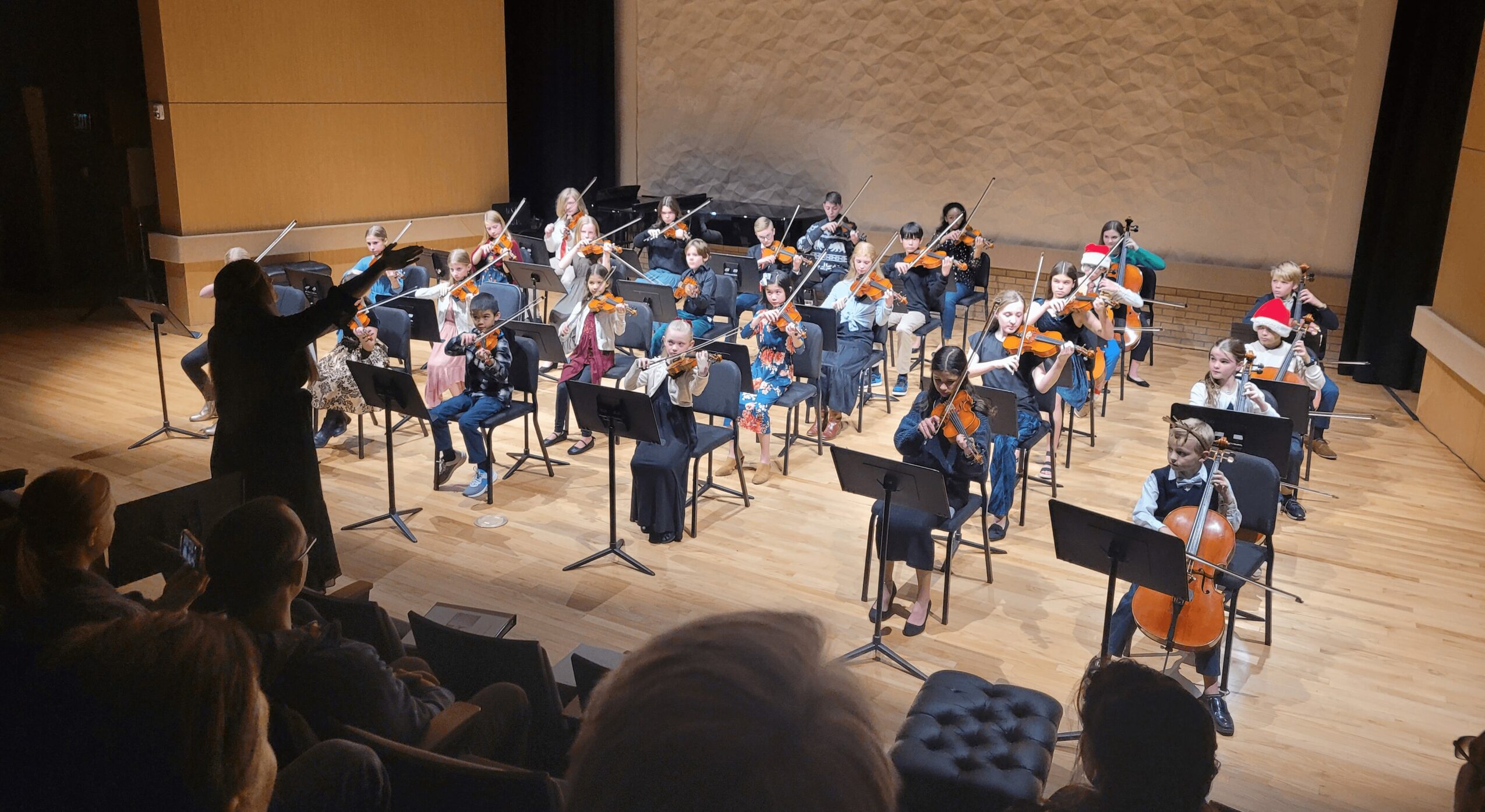 Orchestra of children playing instruments on stage with conductor, audience watching, in a concert hall.