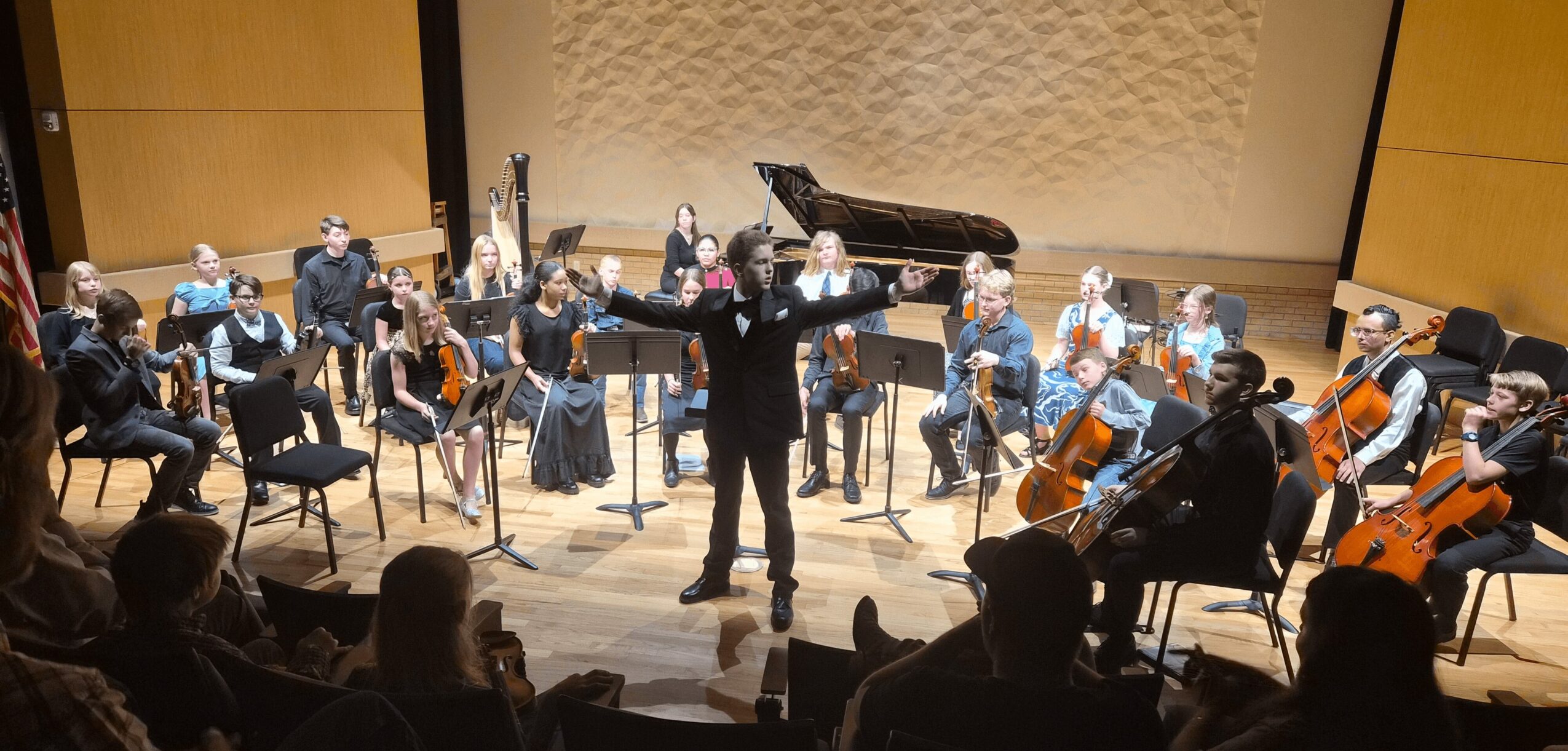 Orchestra performing on stage with conductor, musicians playing string and wind instruments, audience watching, grand piano in background.