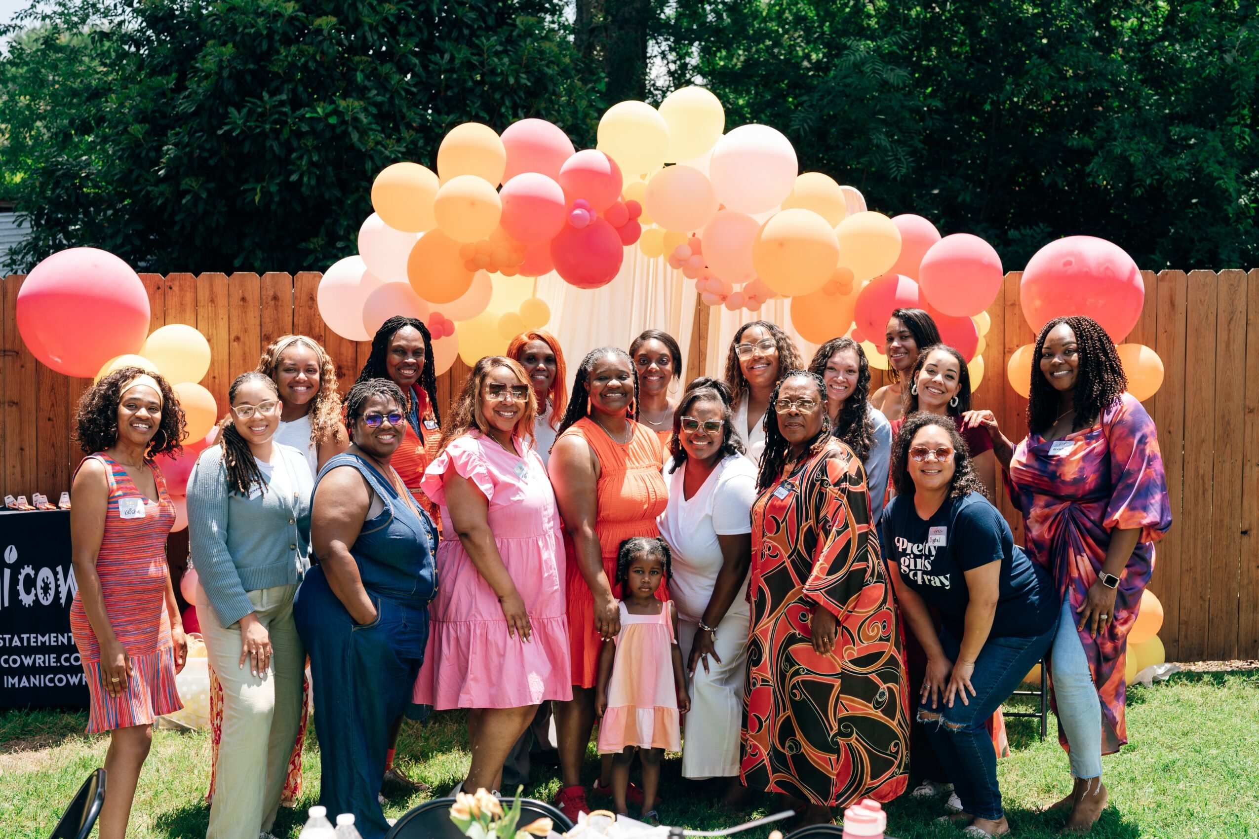 Group of women and children standing outdoors with colorful balloons and a wooden fence in the background.
