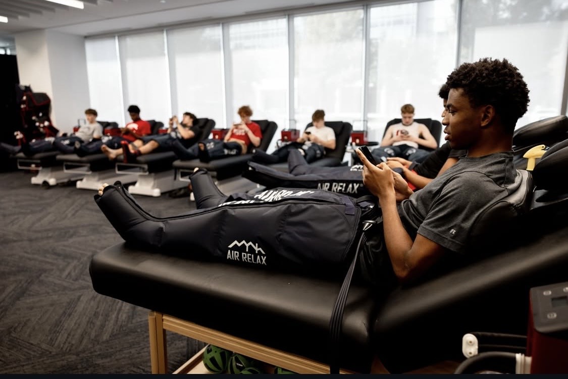 Group of people lying on massage chairs in a room with large windows, some using electronic devices.