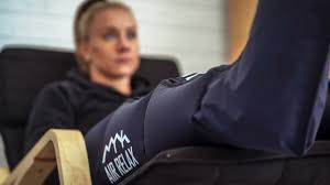 Person sitting on a couch with legs extended, wearing dark pants with a logo, in a room with a woman in the background.