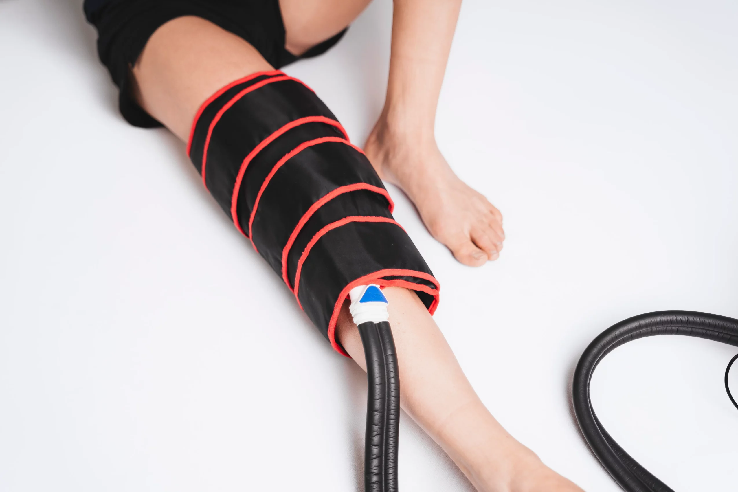 Person with a leg in a black and red compression sleeve, sitting on a white surface, with a black hose nearby.