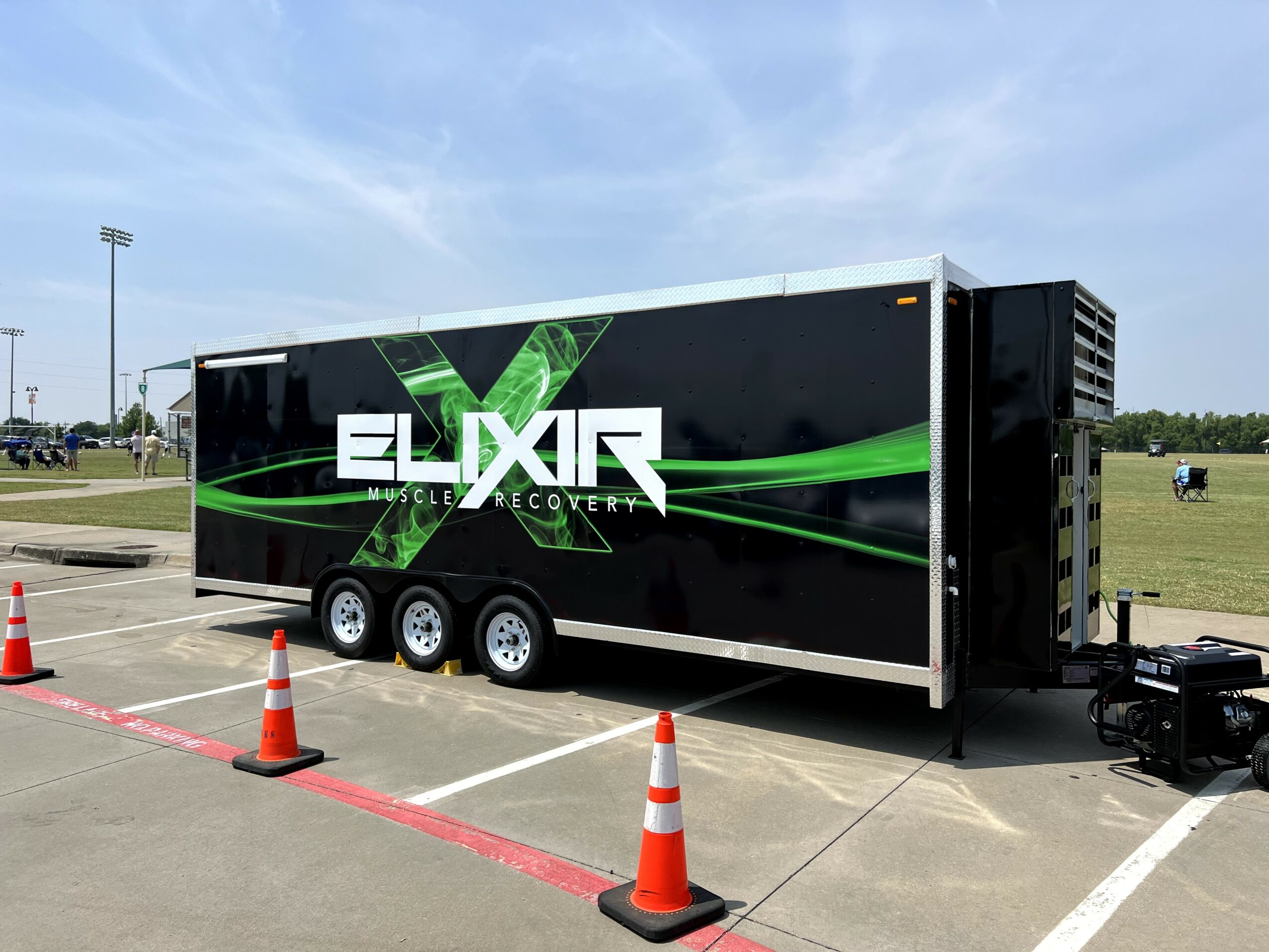 Large black trailer with green and white graphics and the word 'ELIXIR' parked in a lot with orange cones, grassy field, and blue sky.
