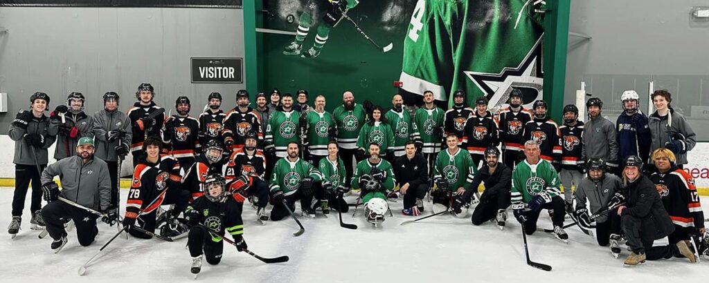 Group of hockey players and coaches on ice rink, some in green jerseys, others in black and red, with a large green banner in background.