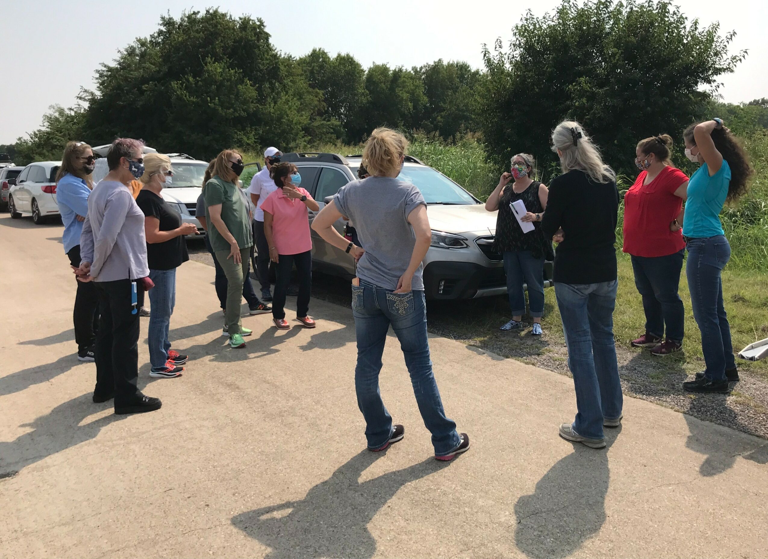 Group of people gathered outdoors near parked cars, listening to a person speaking, with trees and a clear sky in background.