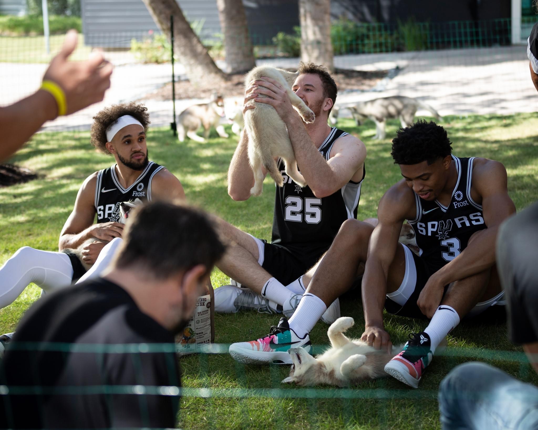Basketball players sitting on grass, one holding a dog, others relaxing outdoors, trees in background, sunny day.