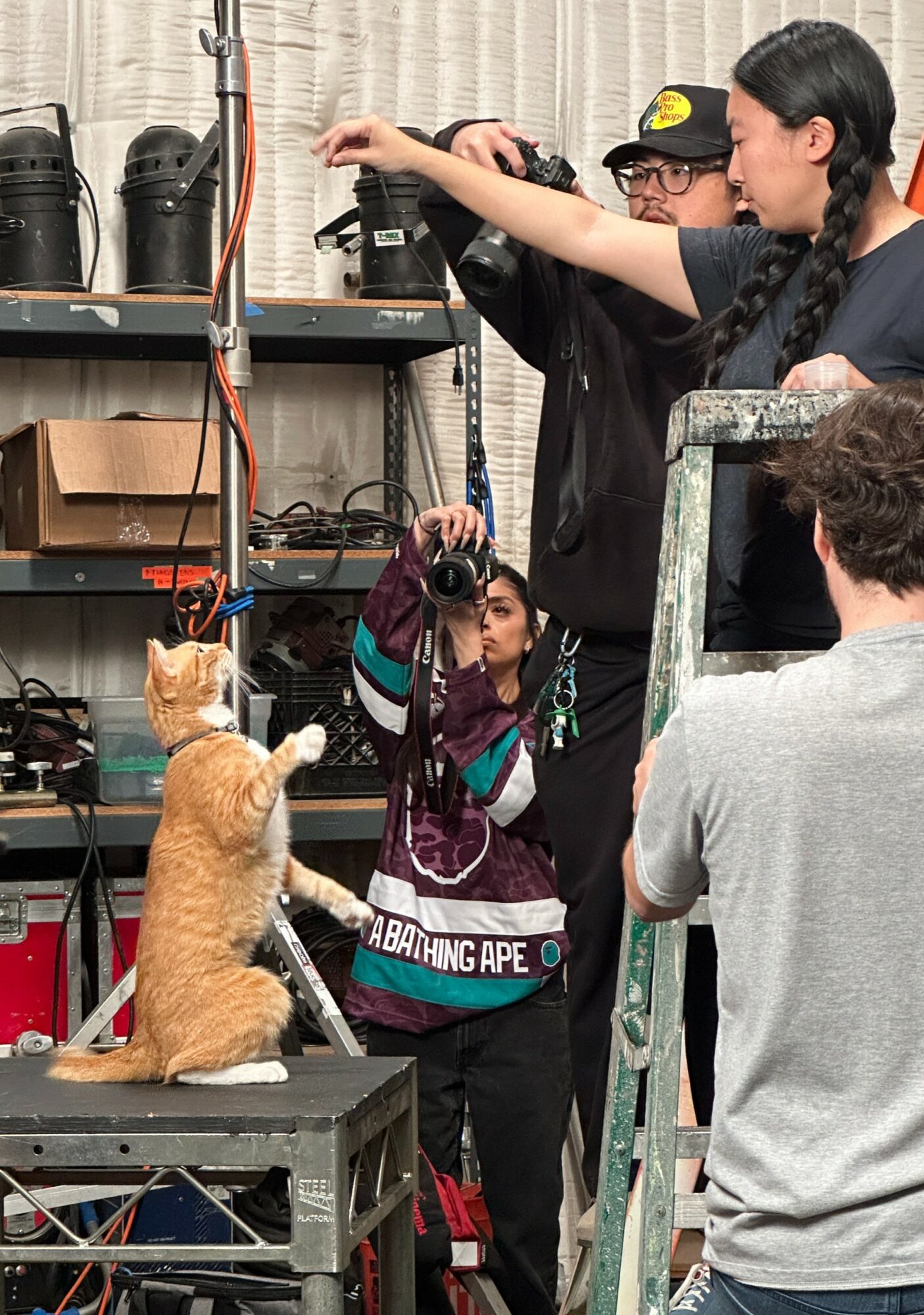Group of people in a workshop, with a woman holding a camera and a person pointing at equipment, and a cat on a table.