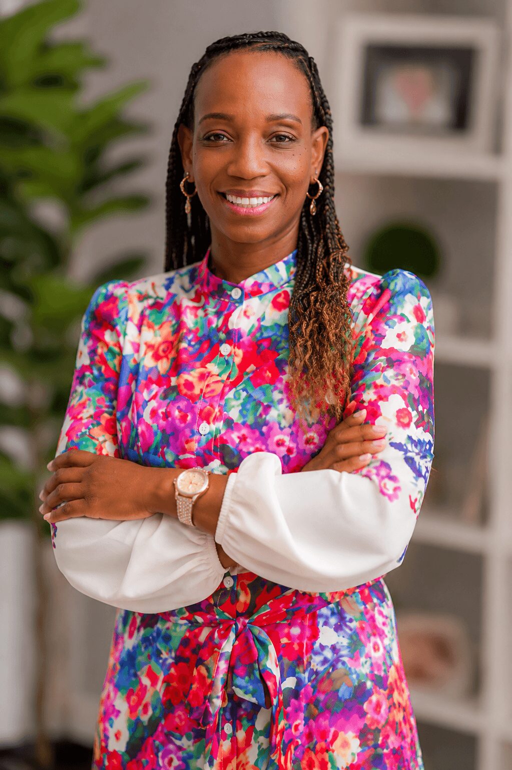 Smiling woman with braided hair wearing a colorful floral dress and white sleeves, standing indoors with shelves and a plant behind her.