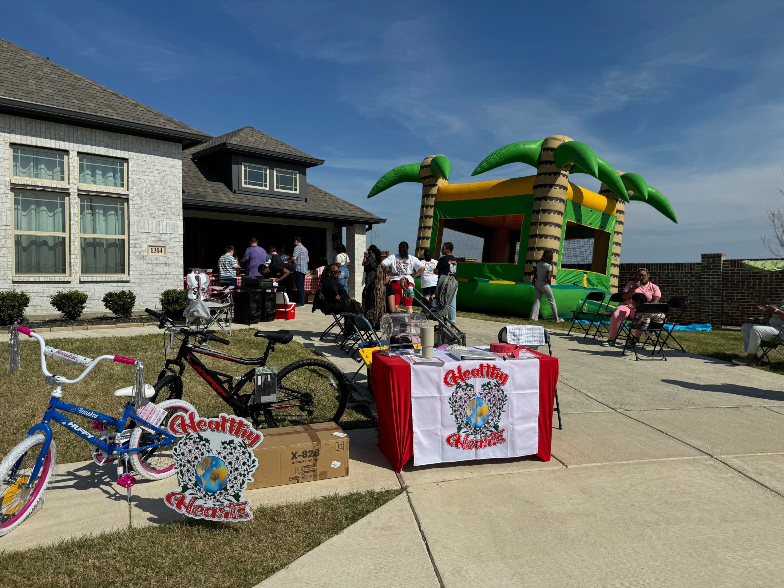 People gather outside a house with a bounce house and tables, children’s bikes, and decorations for a celebration.
