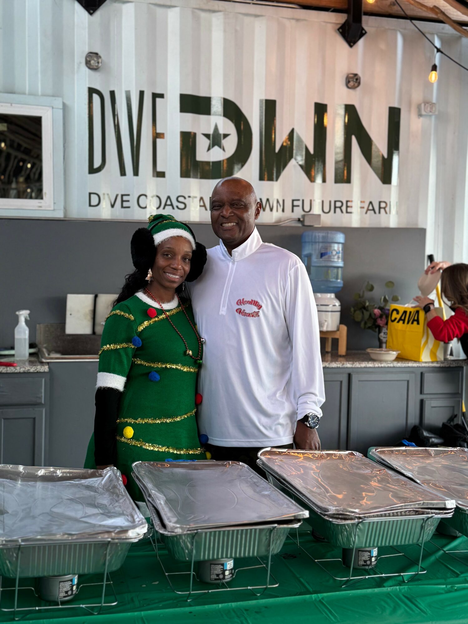 Two people smiling in front of a sign that says 'DIVE DOWN' at a food event, with food trays in front.