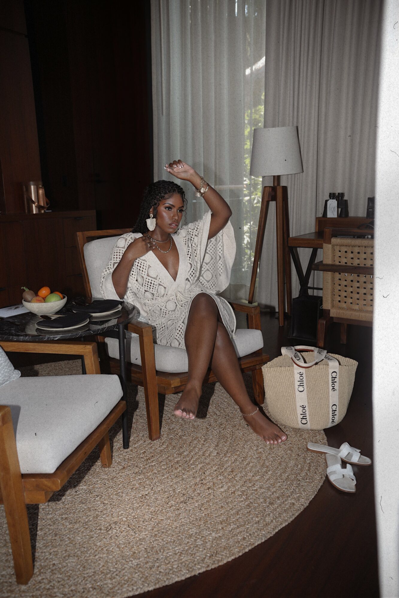 Woman sitting on a chair in a room with curtains, a lamp, and a suitcase, adjusting her hair.