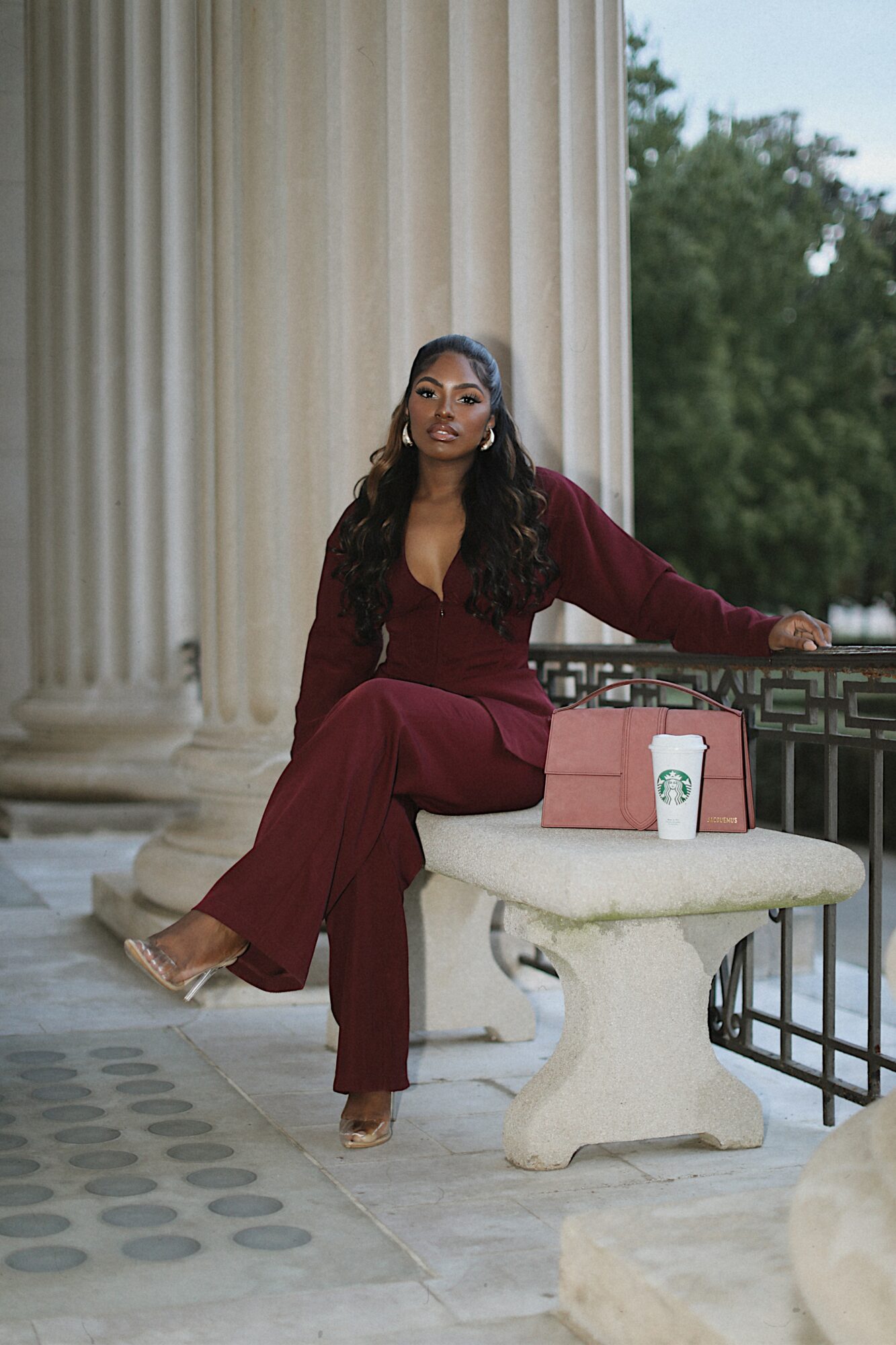 Woman in maroon outfit sitting on a stone bench outdoors, with columns and trees in the background.