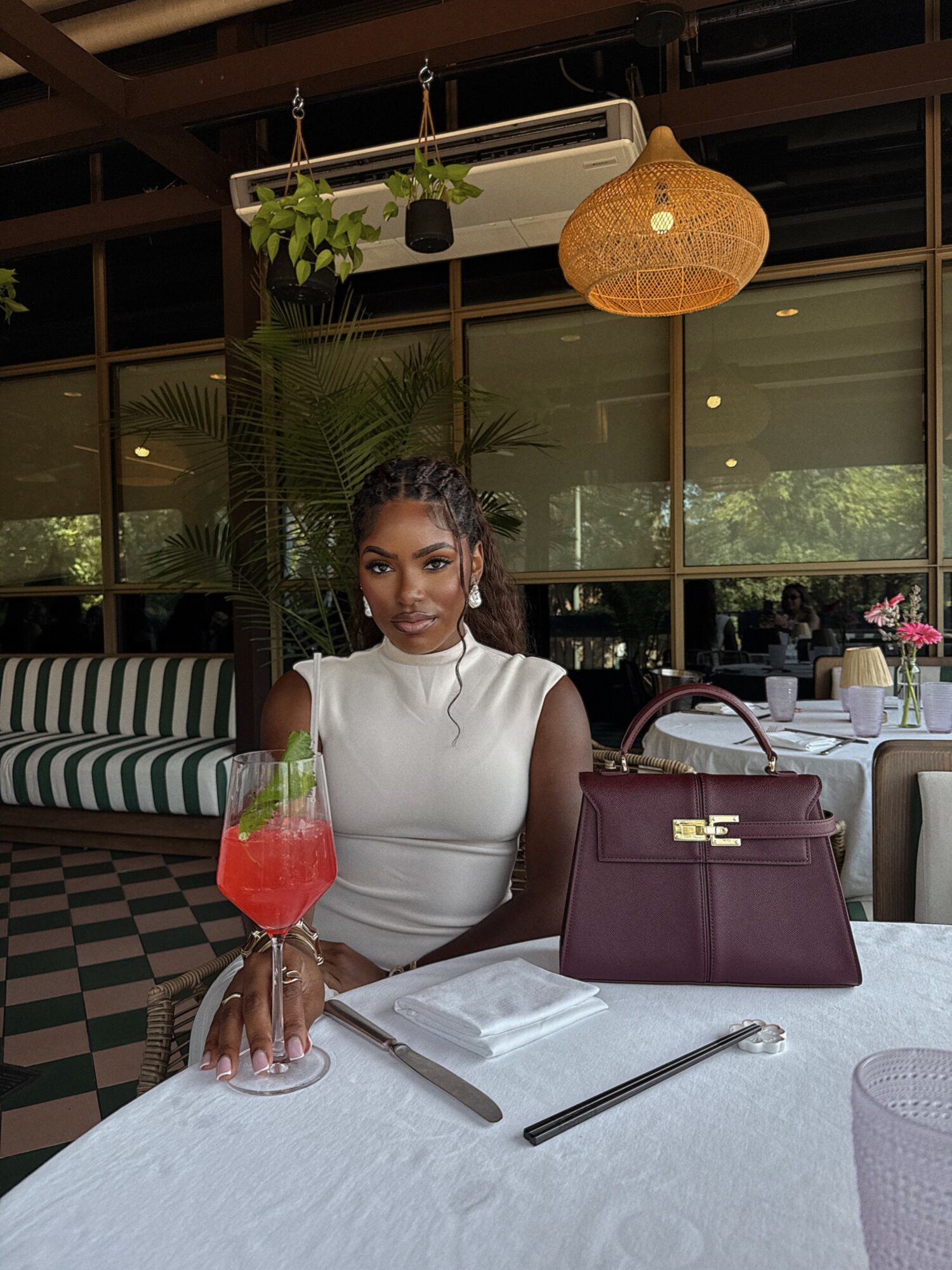 Woman sitting at a table with a pink drink, purple handbag, and utensils in a restaurant with hanging plants and wicker lamp.
