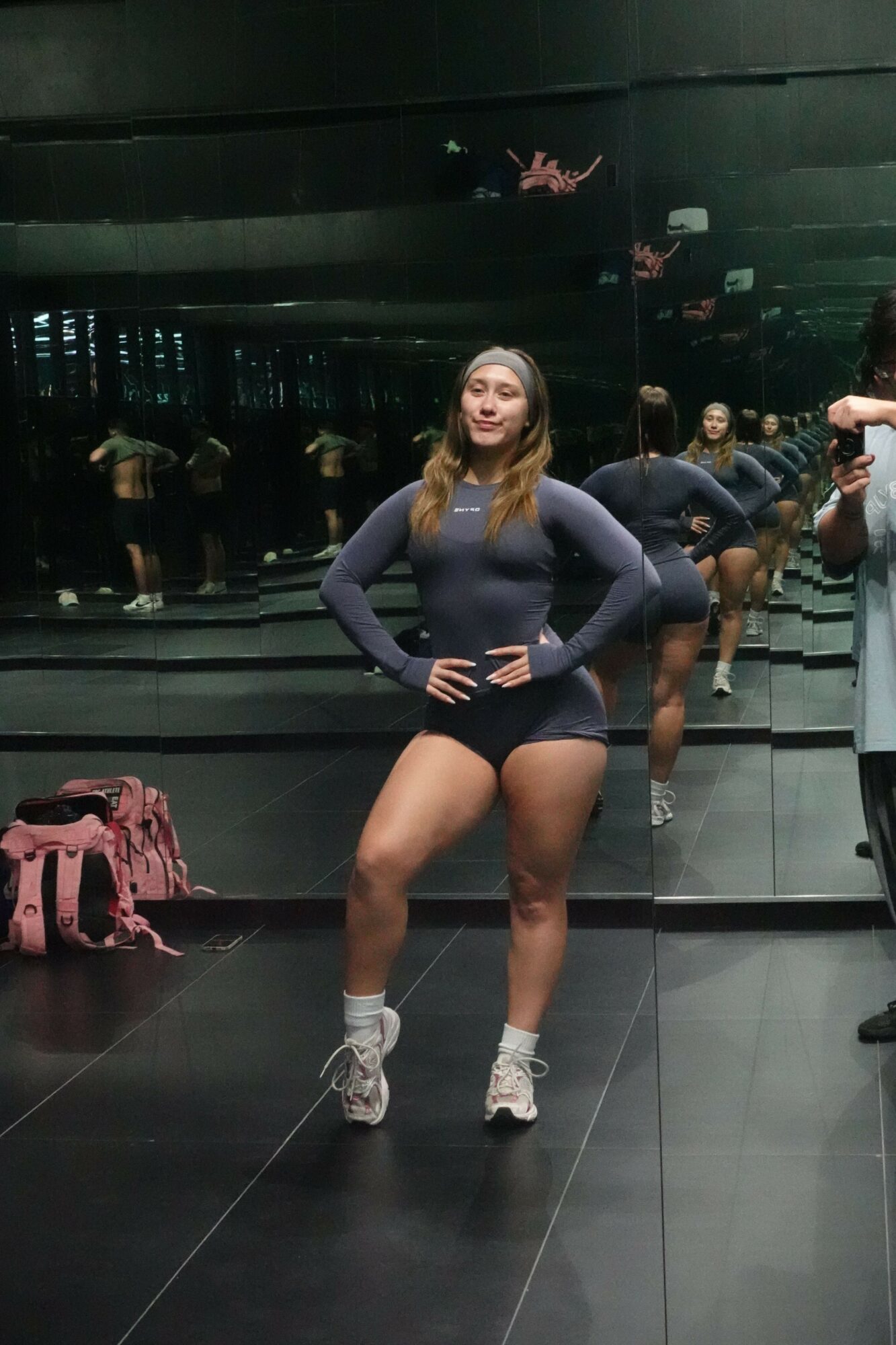 A woman in workout clothes takes a mirror selfie in a gym, with other people and gym equipment in the background.