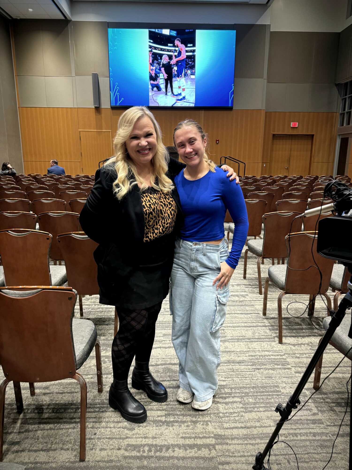 Two women standing together in an auditorium with empty chairs, a large screen behind them displaying a photo, and a camera on a tripod nearby.