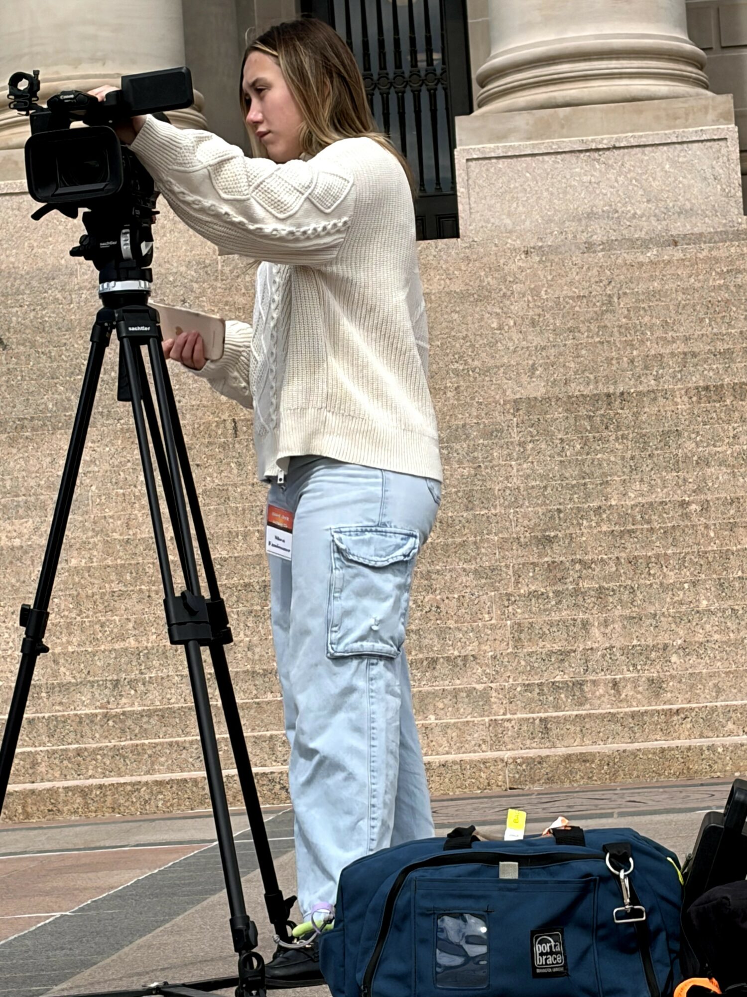Young woman operating a camera on a tripod outside a building with stone steps and columns.