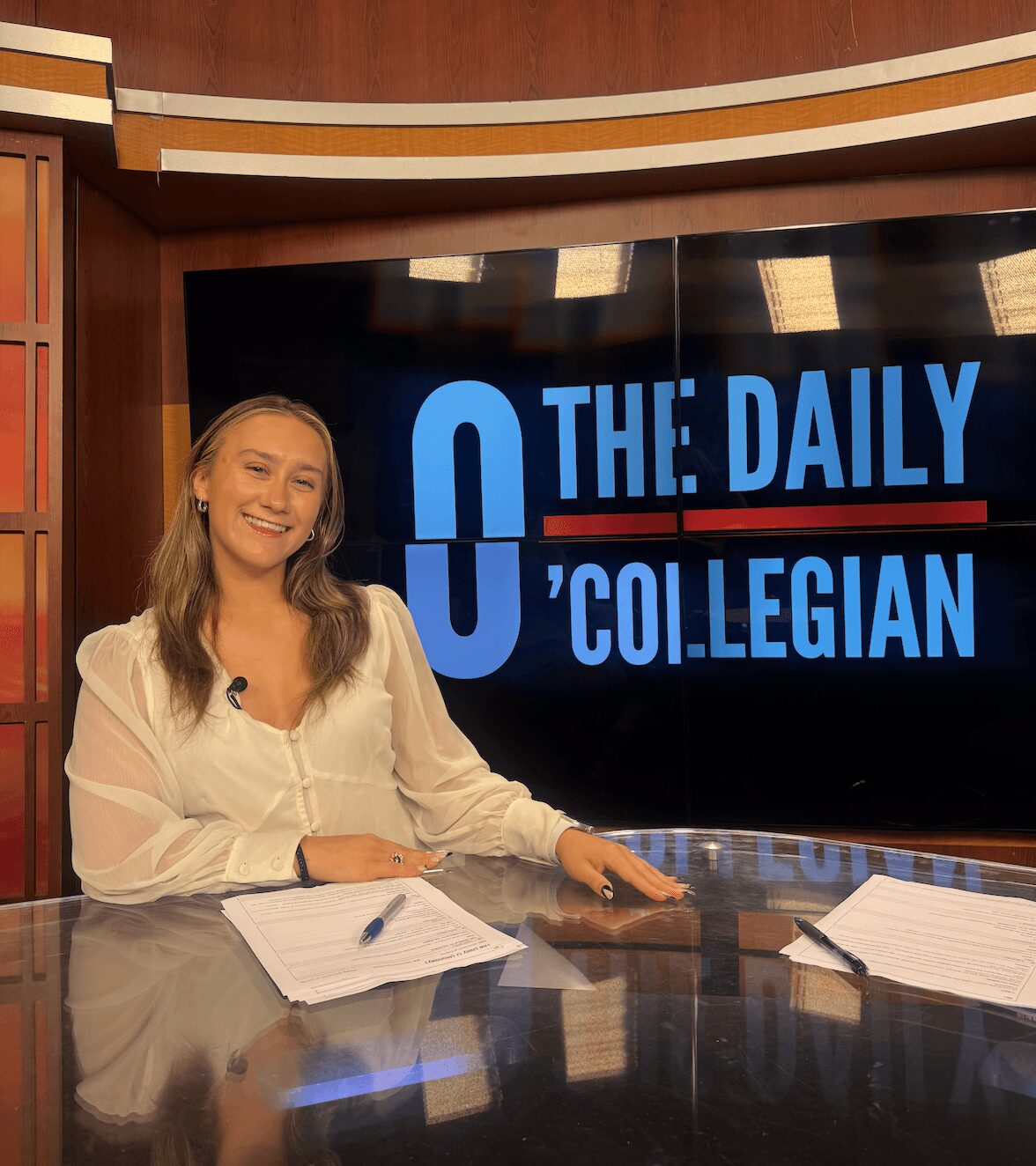 Woman with long hair smiling at news desk with papers, behind a large screen displaying 'The Daily Collegian' logo.