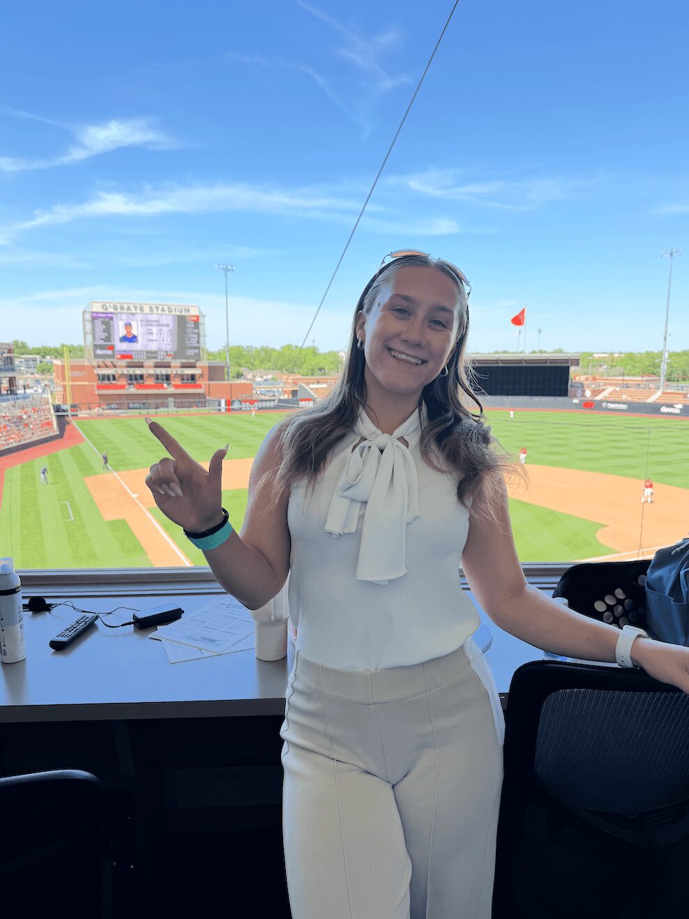 Smiling woman in white outfit standing in a stadium press box with a baseball field in the background.