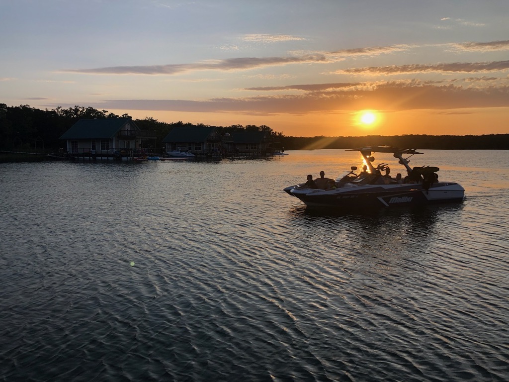 Boat with people on water during sunset, with a distant shoreline and cloudy sky.
