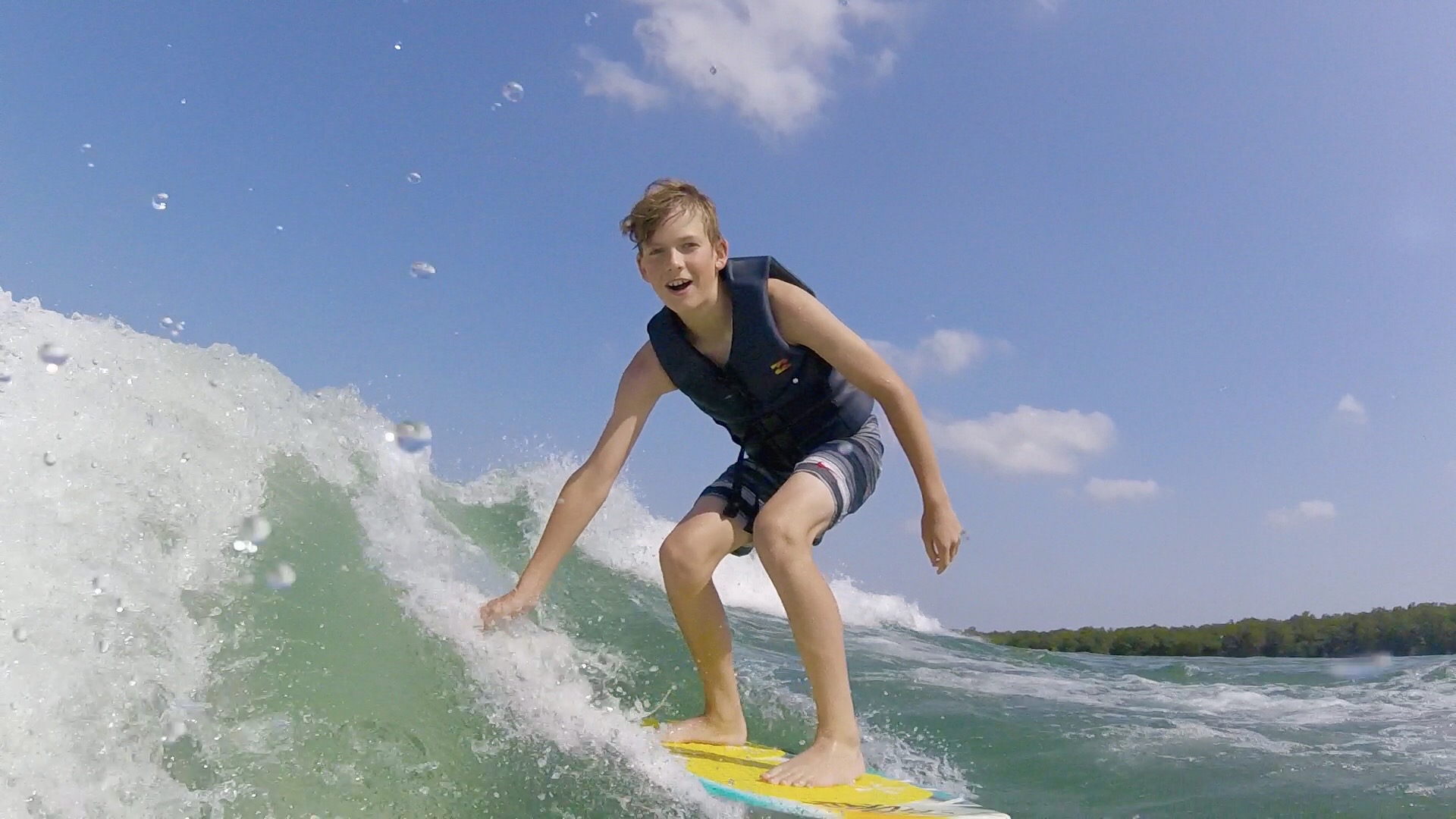 Child surfing on a wave with a blue sky and distant shoreline in the background.