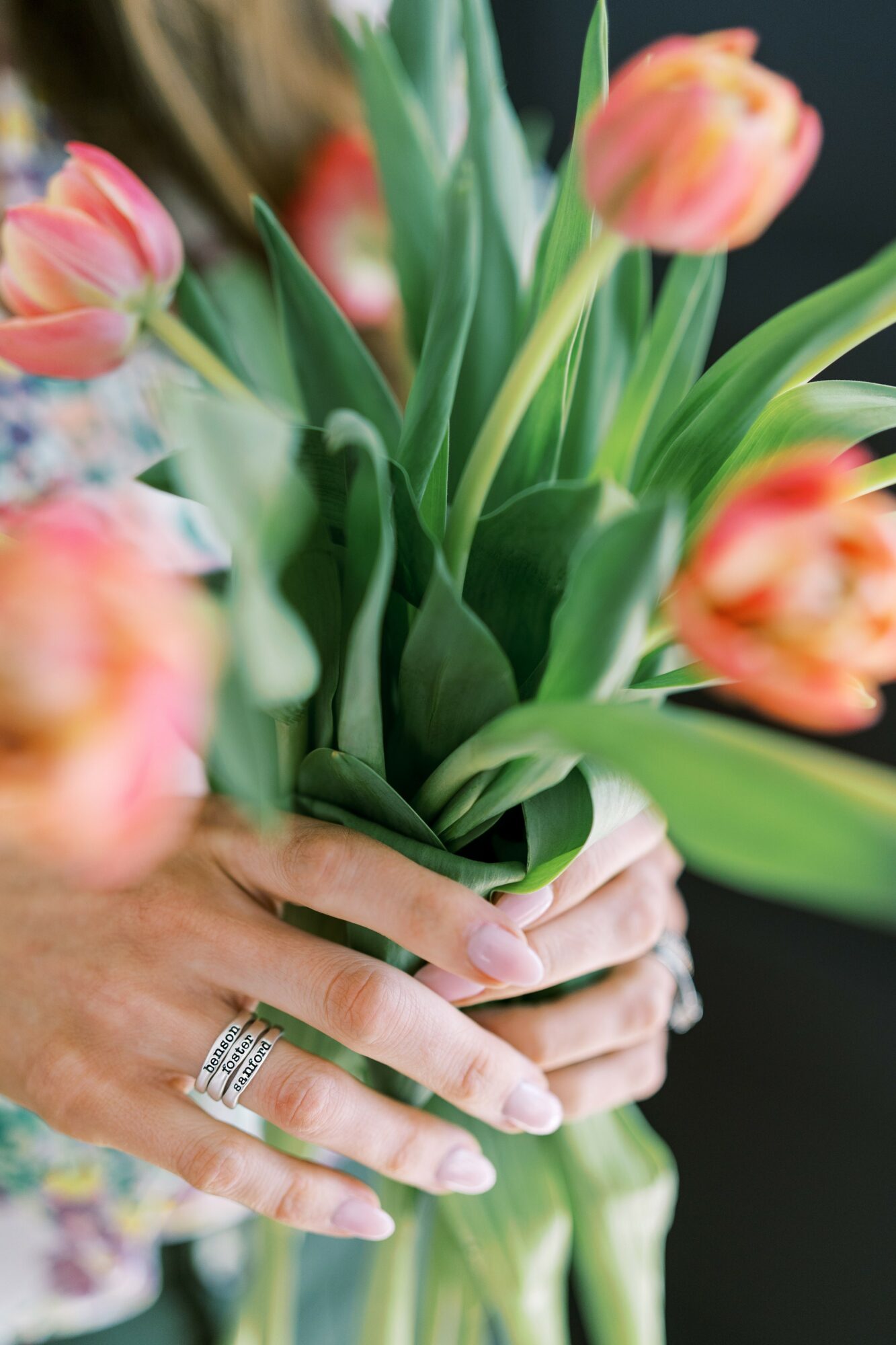 Person holding a bouquet of pink and orange tulips with green leaves, wearing rings on fingers.