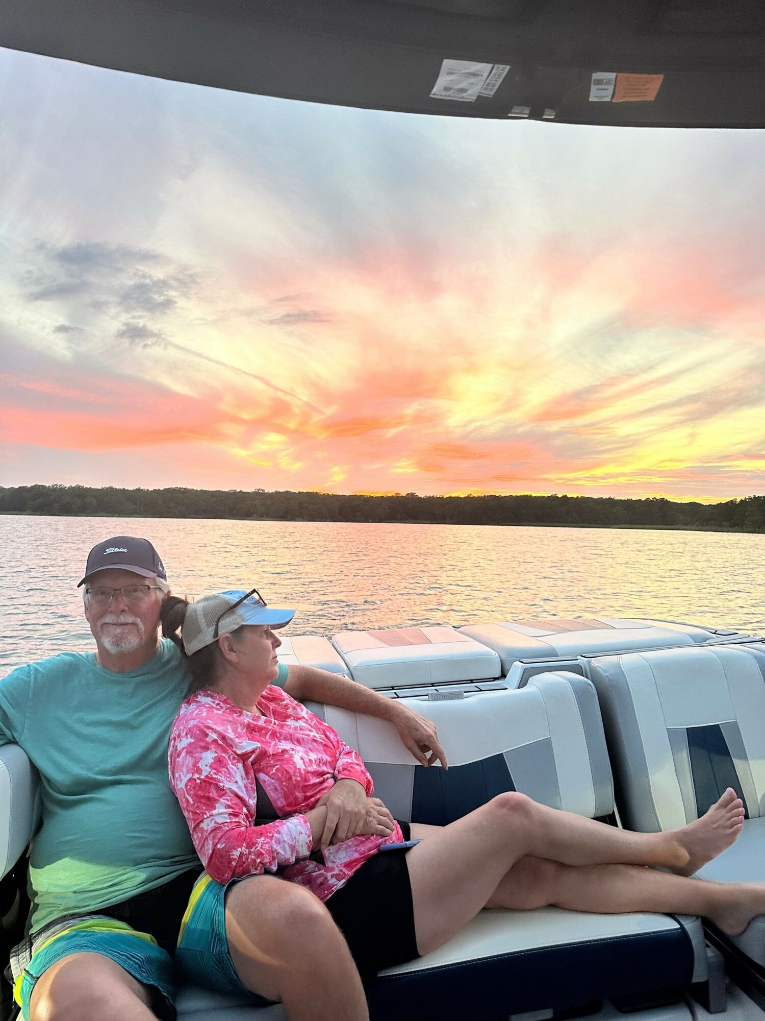 Person lying on a boat with legs crossed, wearing a pink shirt, at sunset over water.