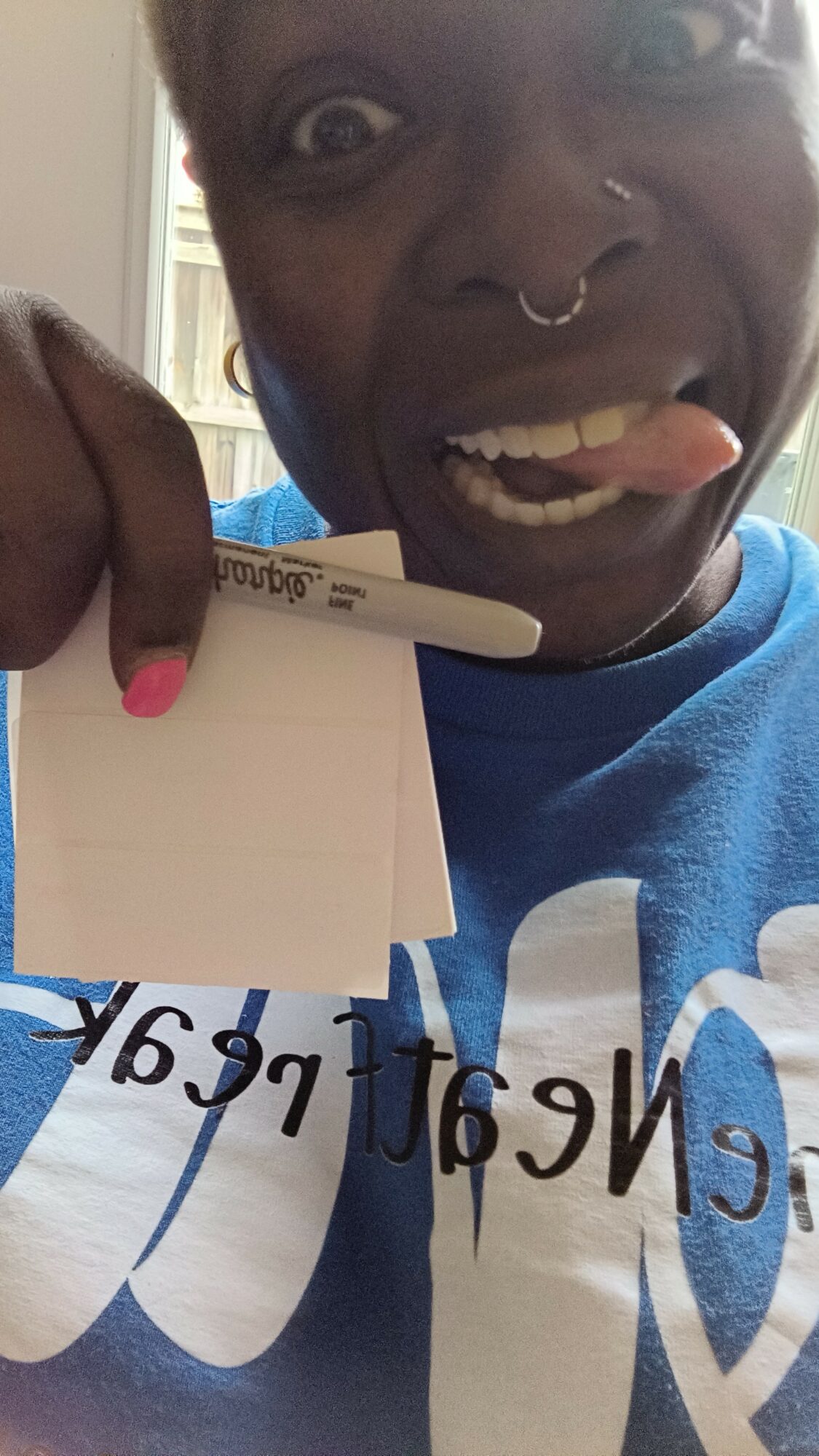 Person with a septum piercing and tongue out holding a whiteboard with handwritten text, wearing a blue shirt.