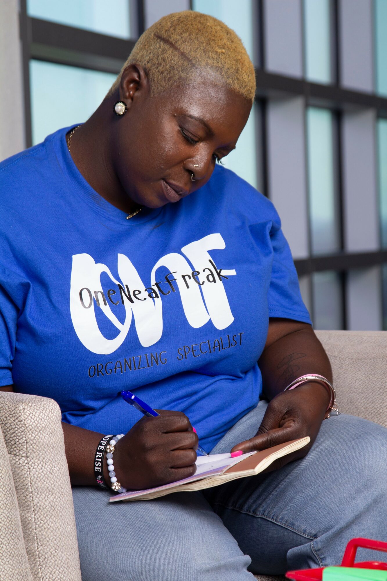 Woman with short blonde hair writing in a notebook, wearing a blue T-shirt, sitting indoors.